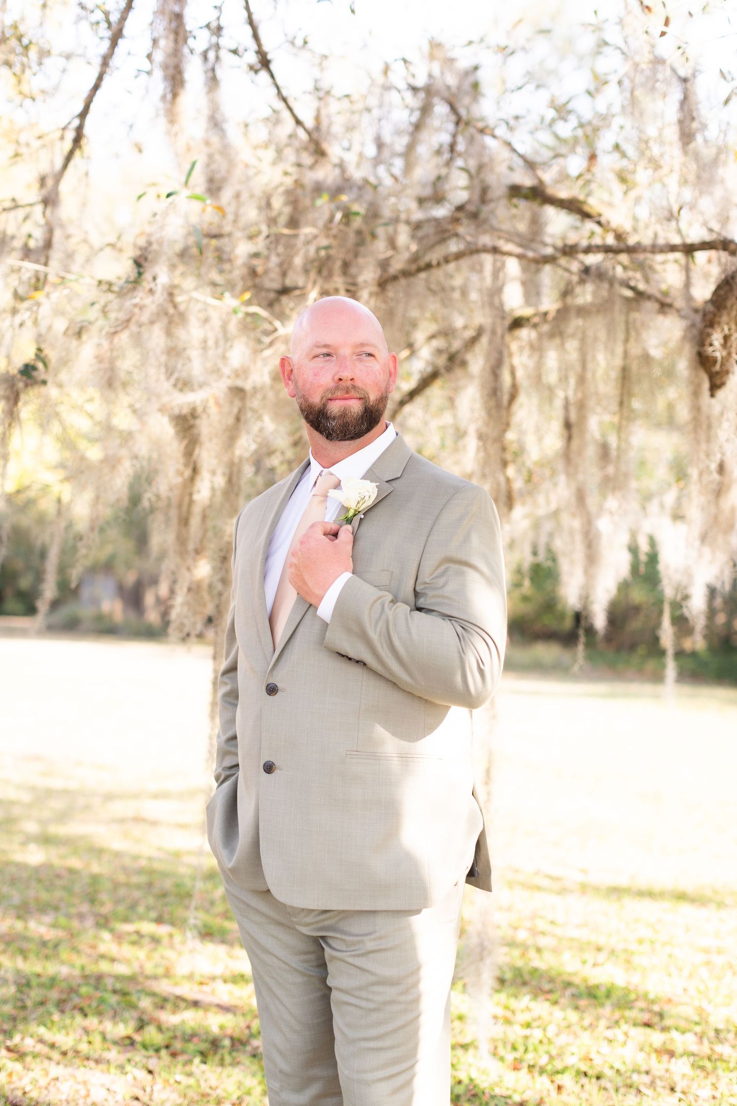 Groom in light gray suit poses confidently against backdrop of moss-covered trees.