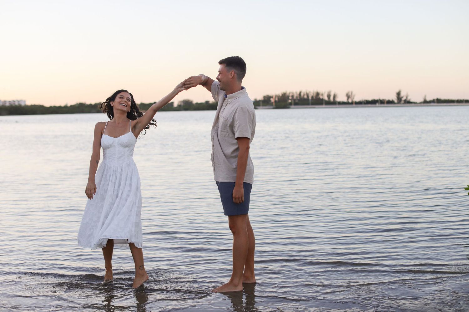 Couple dancing barefoot in shallow water at sunset, man spinning woman in white dress on a serene lake shore.
