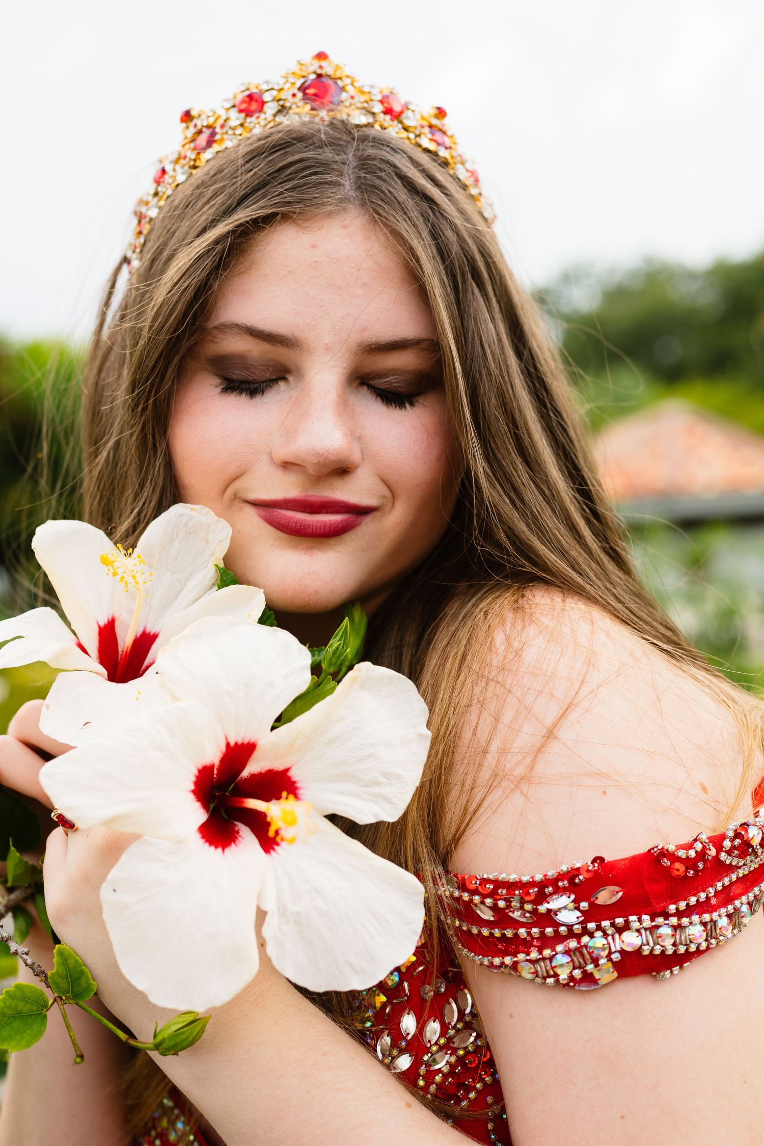 White hibiscus flowers frame a red formal dress with beaded details and floral crown.