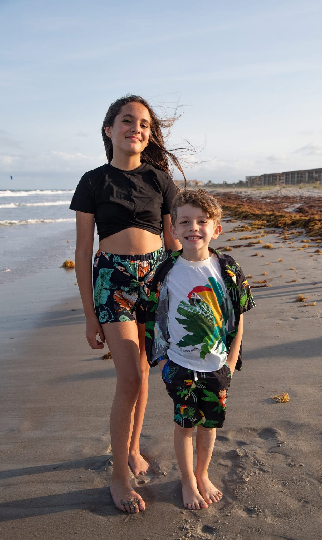 Two people in matching tropical print outfits stand together on a beach at sunset.