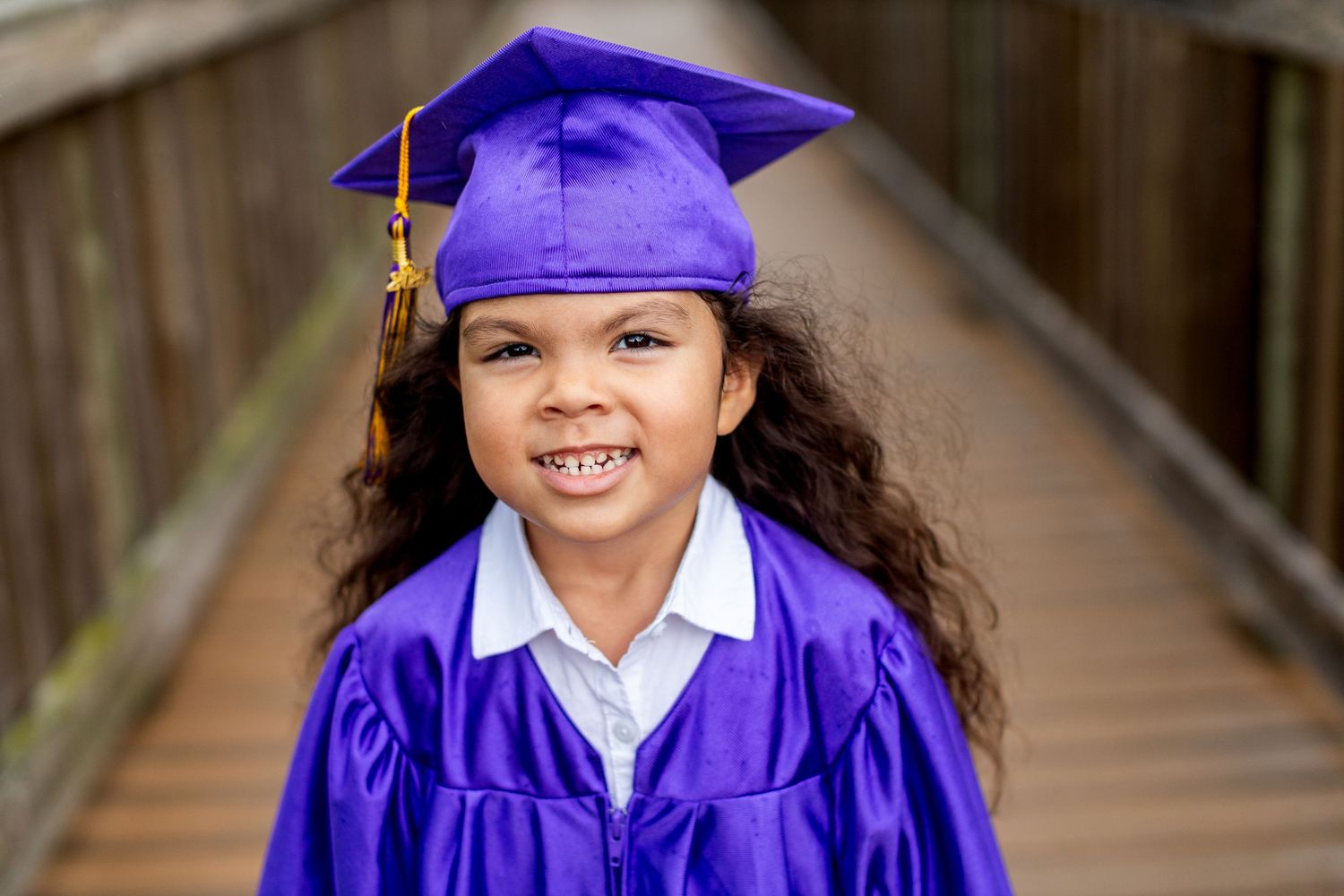 Graduate wearing purple academic regalia poses cheerfully on wooden walkway.