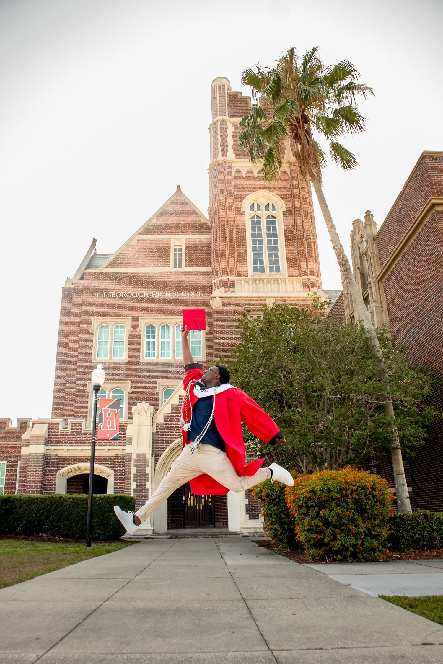 Graduates in red robes perform acrobatic jumps and poses on sidewalk near church.