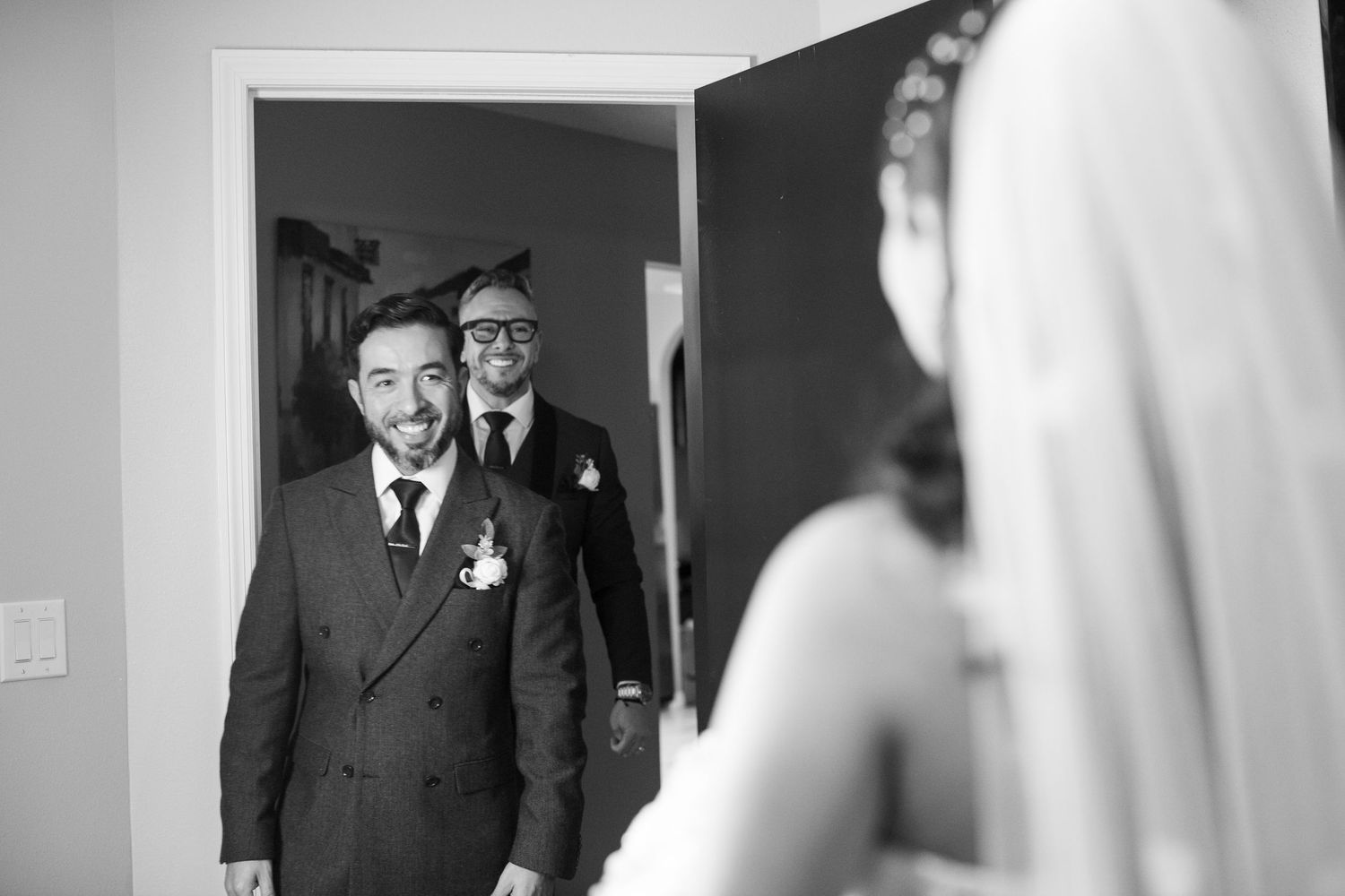 Black and white photo of a groom smiling as he sees his bride for the first time, with a friend behind him.