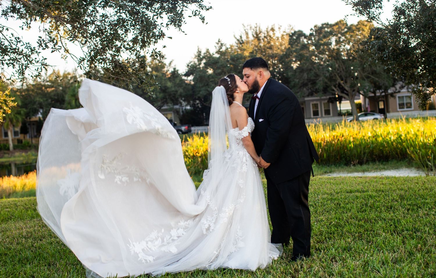 Bride and groom sharing a romantic kiss outdoors, with flowing white veil billowing in the breeze at golden hour.