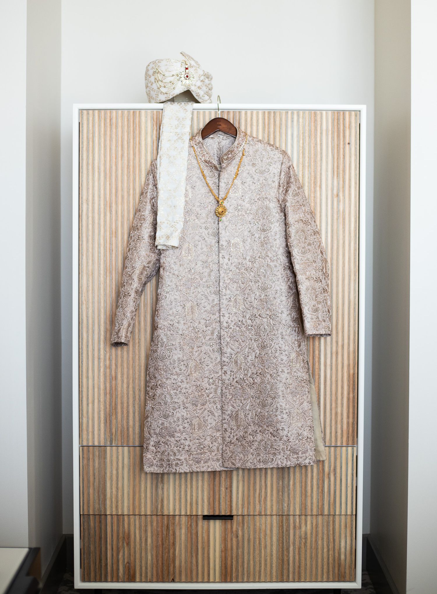 An elegant ivory sherwani and turban displayed on a wooden wardrobe against a striped wall.