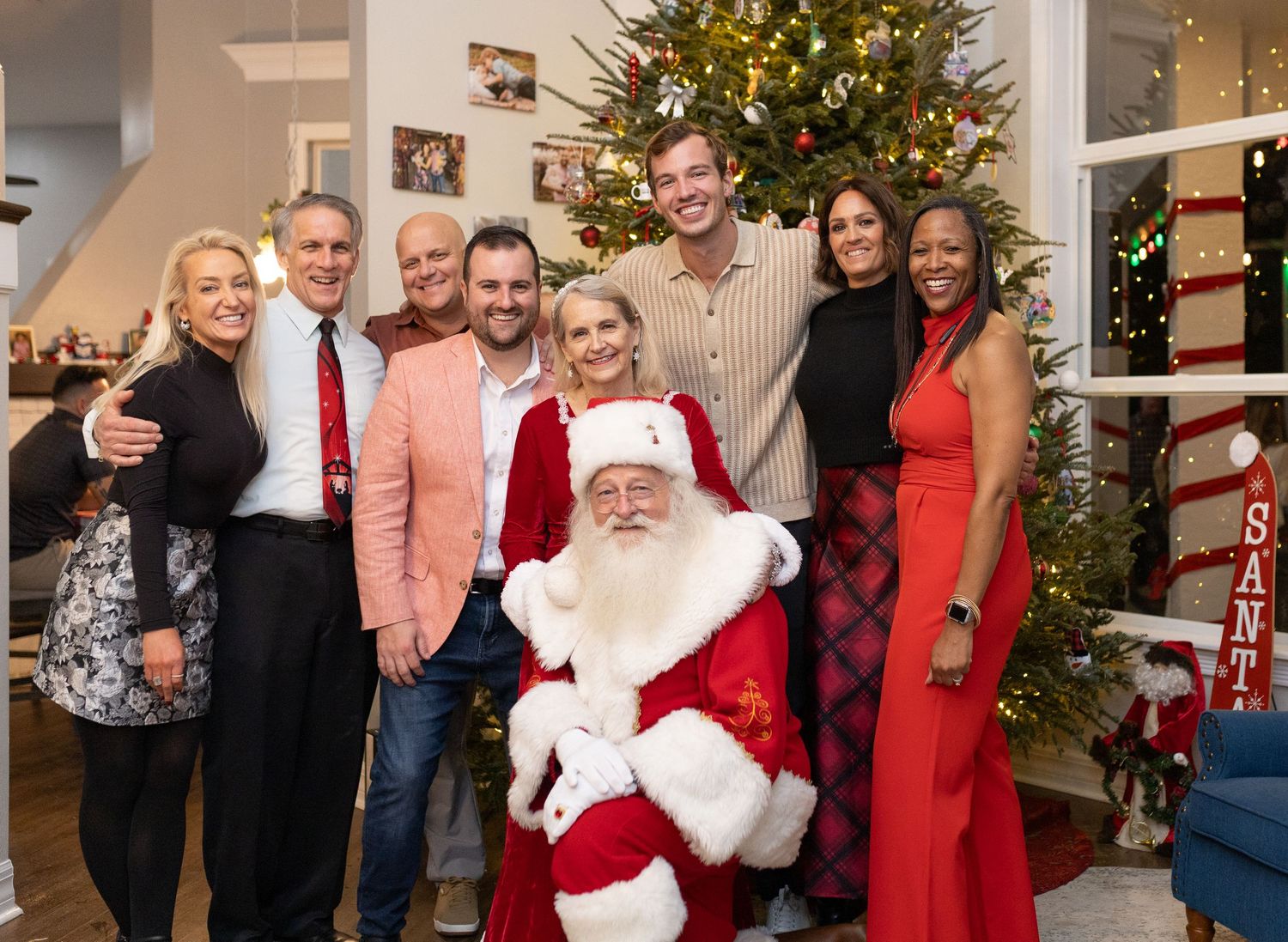 A festive holiday gathering with a decorated Christmas tree and people in formal attire in a bright interior space.