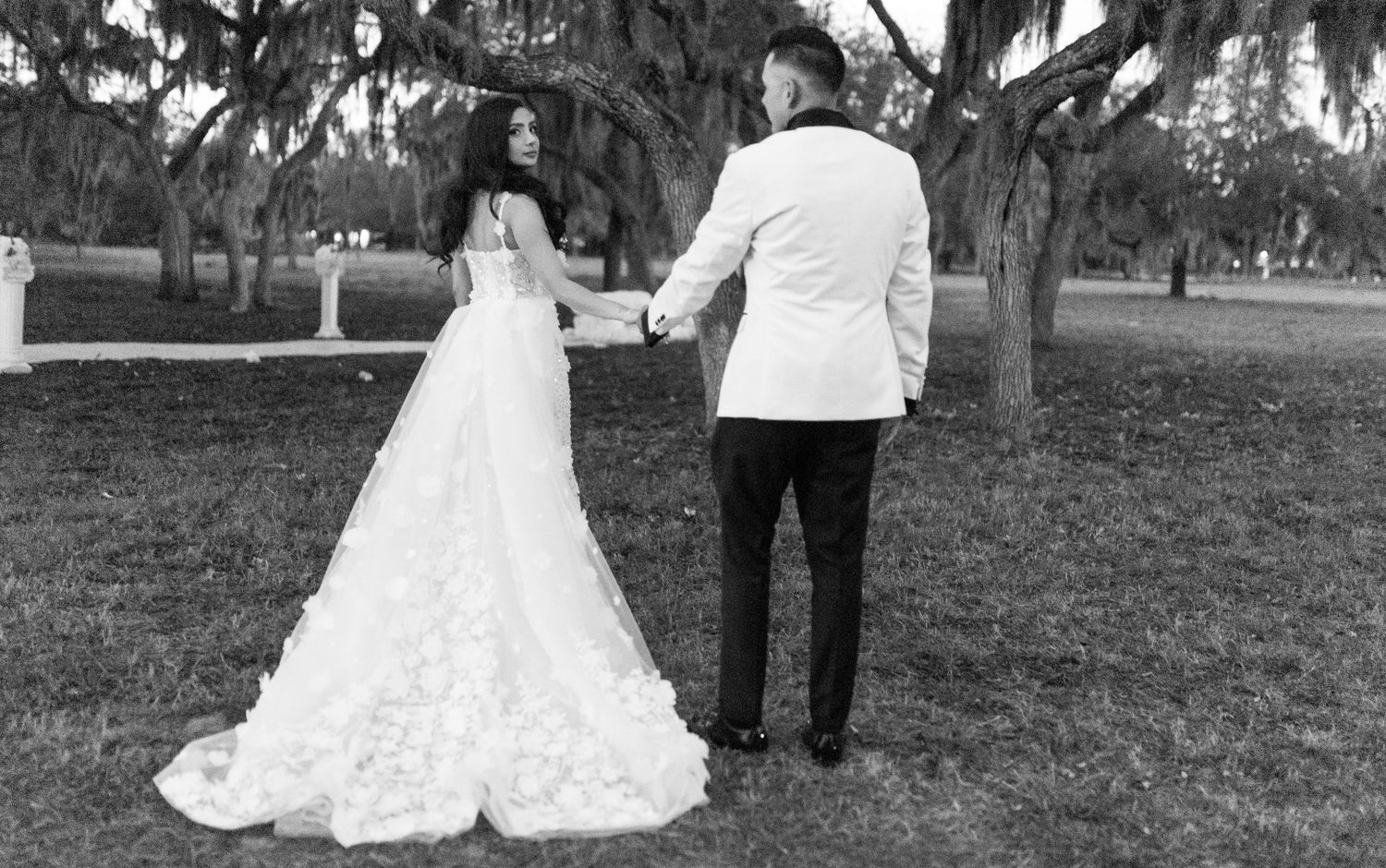 A couple in wedding attire stands together on a lawn with Spanish moss trees in the background.