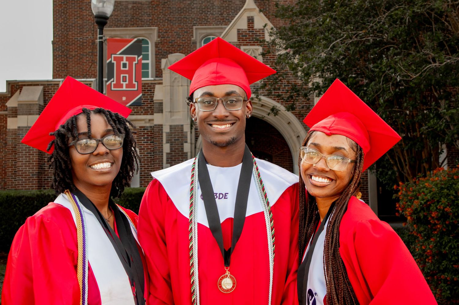Three graduates in red caps and gowns smile together outside brick school building.