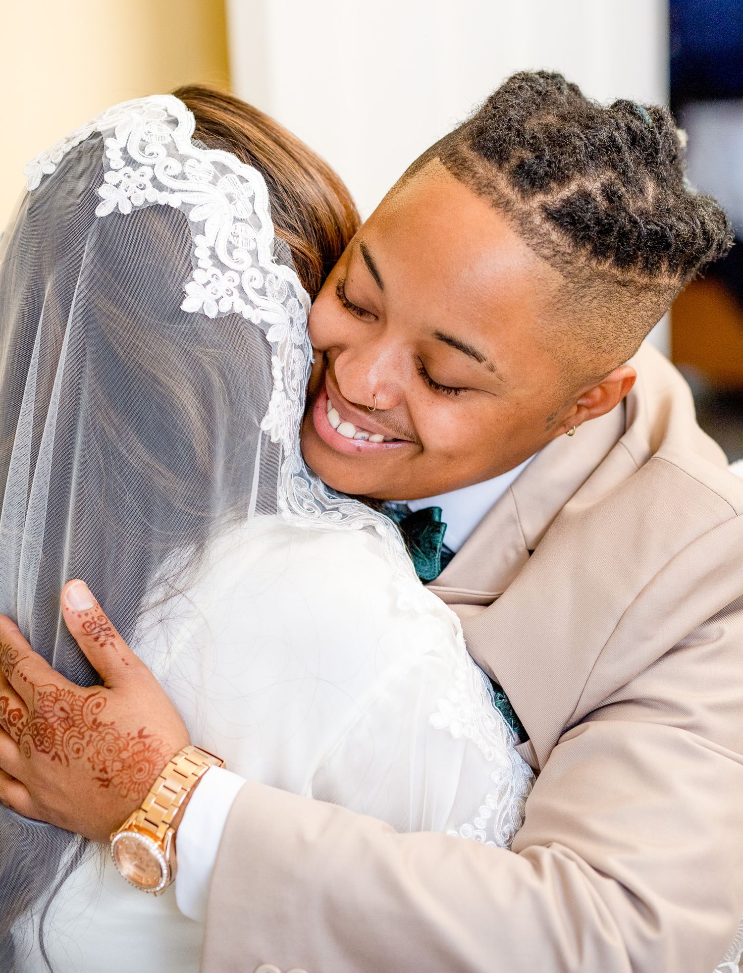 Two people in wedding attire share a joyful embrace, one wearing a white gown with veil and the other in a tan suit.