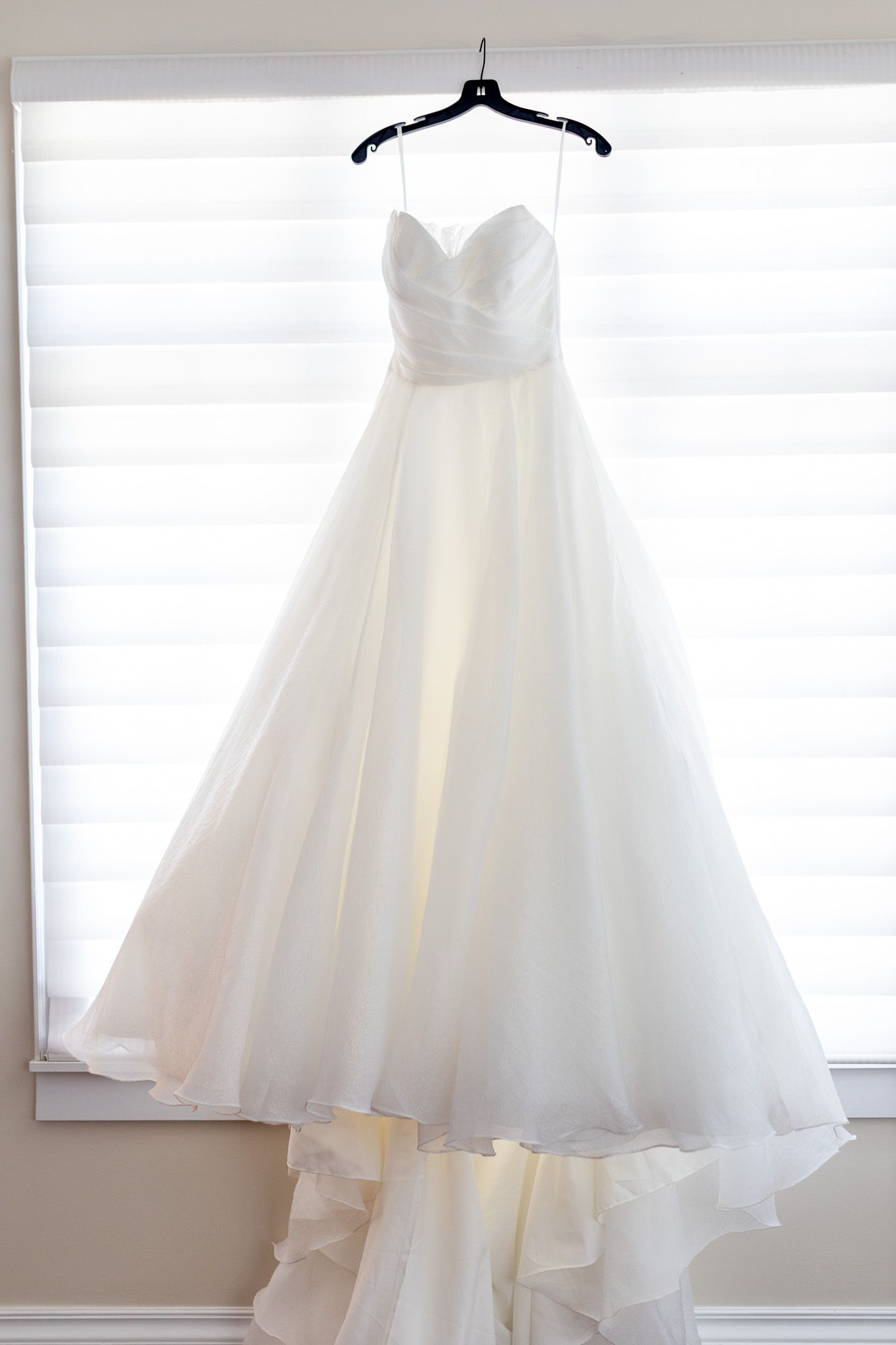 Elegant white strapless ball gown wedding dress hanging on a black hanger in front of a bright window.