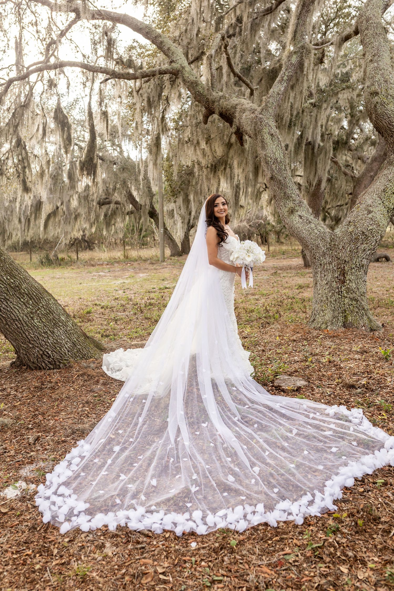 A dramatic cathedral-length lace veil spreads across fallen leaves under moss-draped trees.