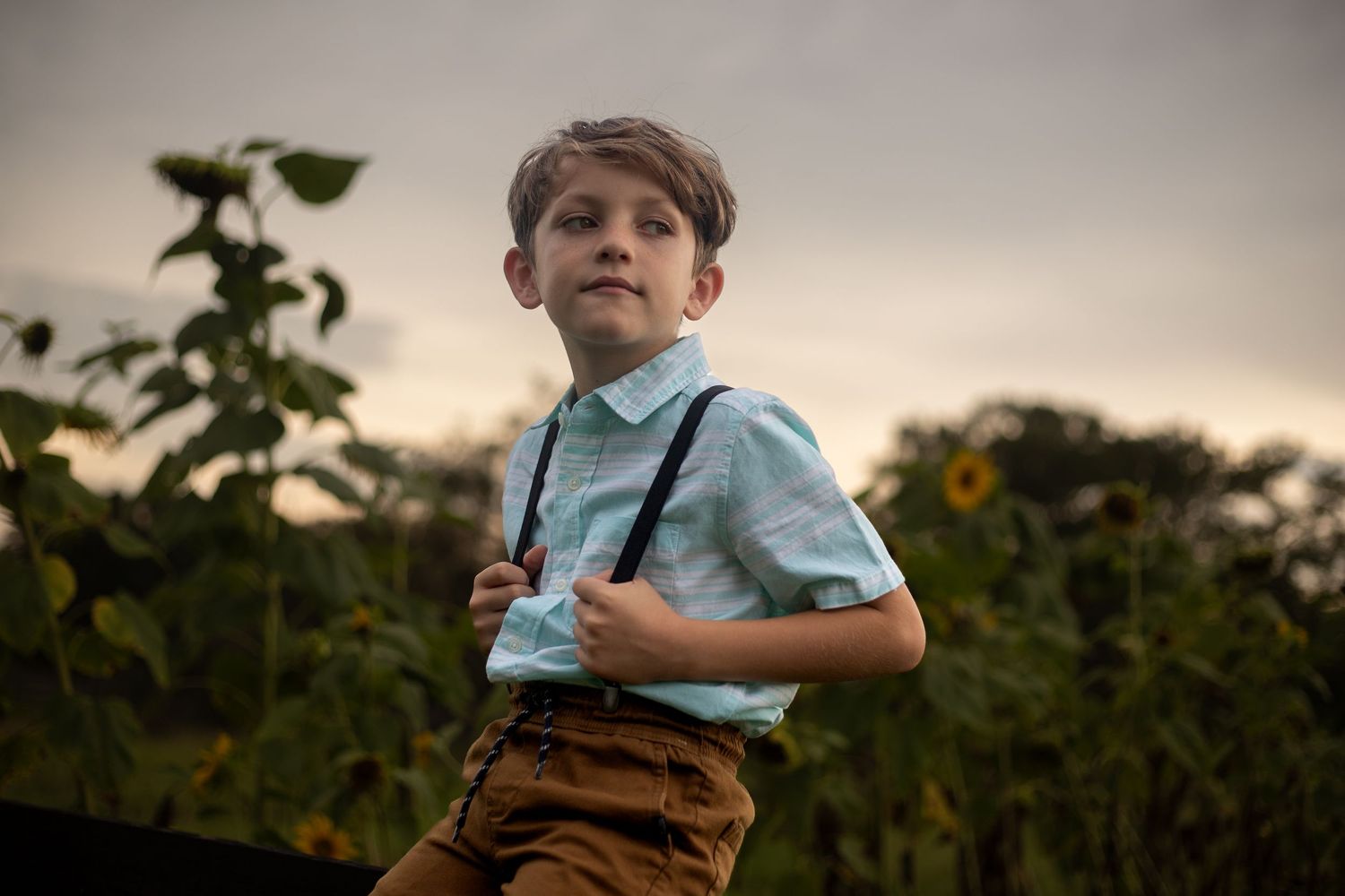 Young child in vintage-inspired outfit with brown shorts and suspenders plays in outdoor garden setting.