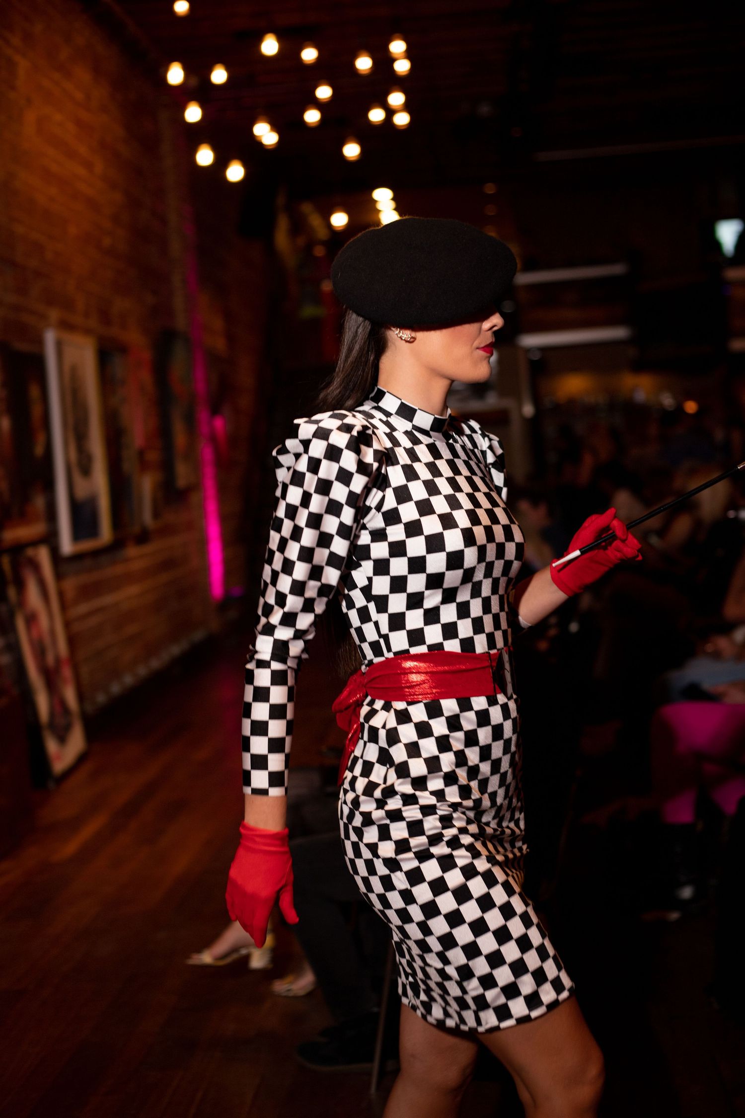 Figure wearing black and white checkered dress with red gloves in dimly lit venue.