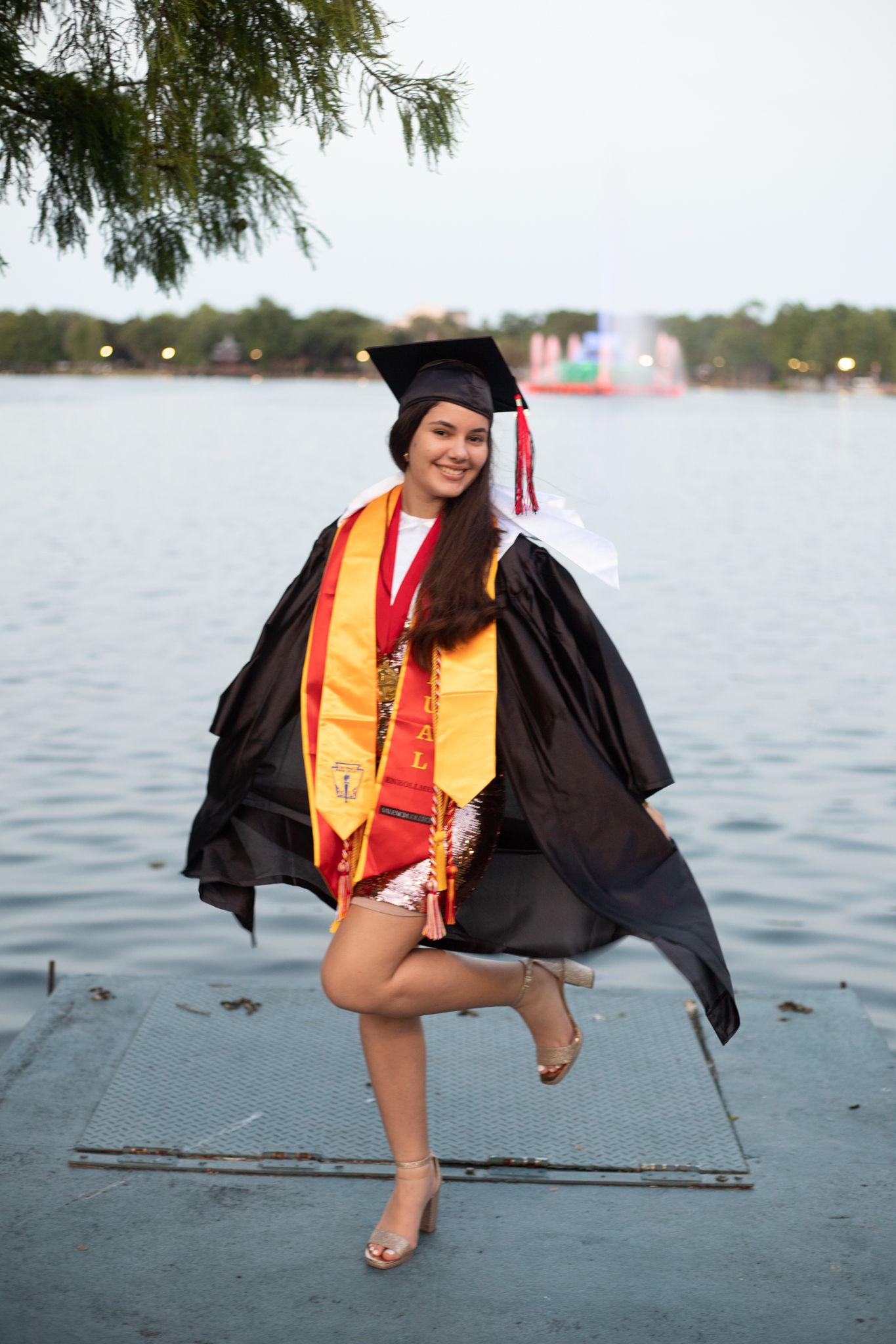 Graduate in black cap and gown with gold stole poses playfully by a lake during sunset celebration.