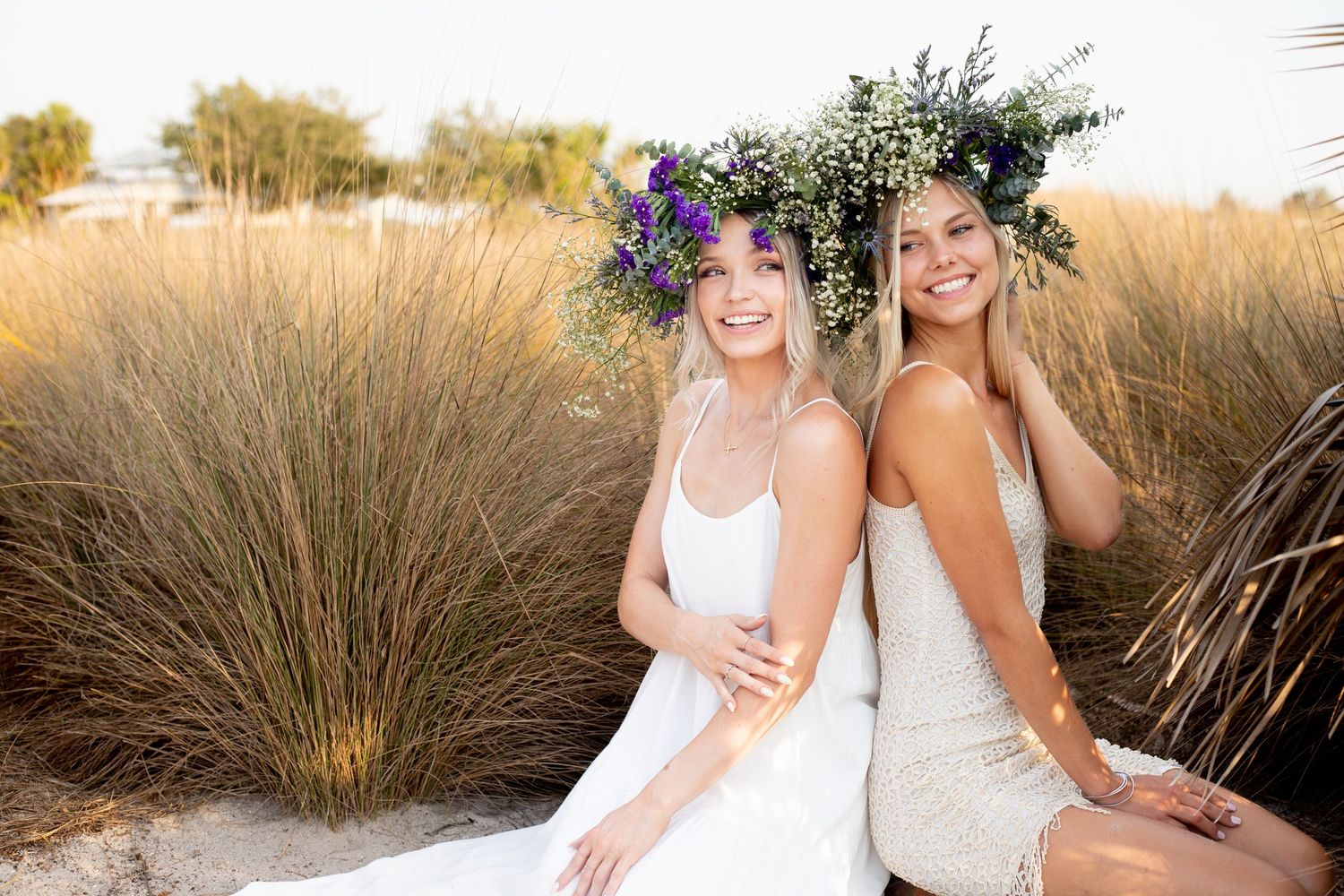 Two women in white dresses wearing floral crowns sitting in tall grass.