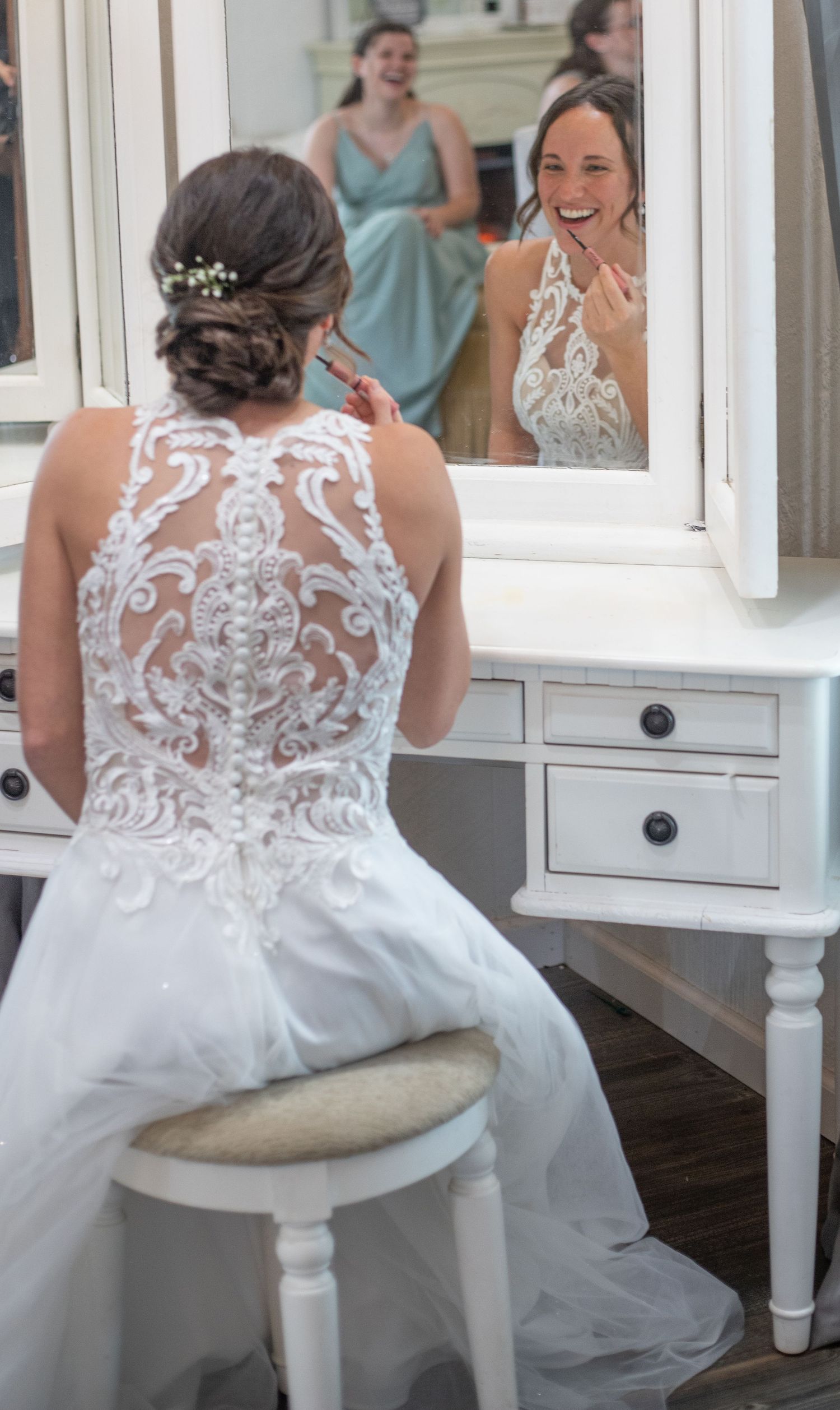 Bride in lace wedding dress sits at vanity mirror, smiling at her reflection while bridesmaid looks on.