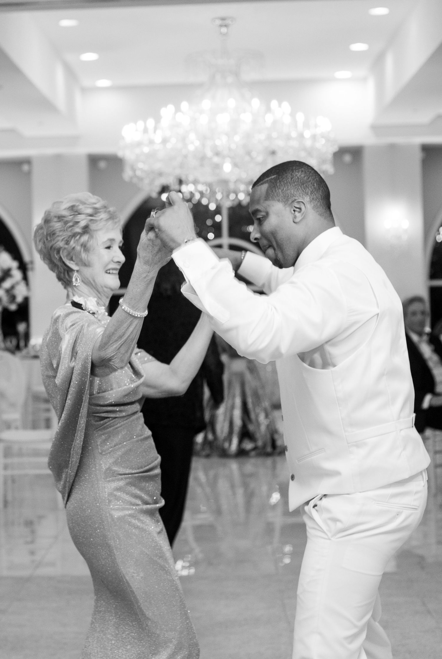 Elderly woman and man dancing joyfully at elegant wedding reception under chandelier, black and white photo.