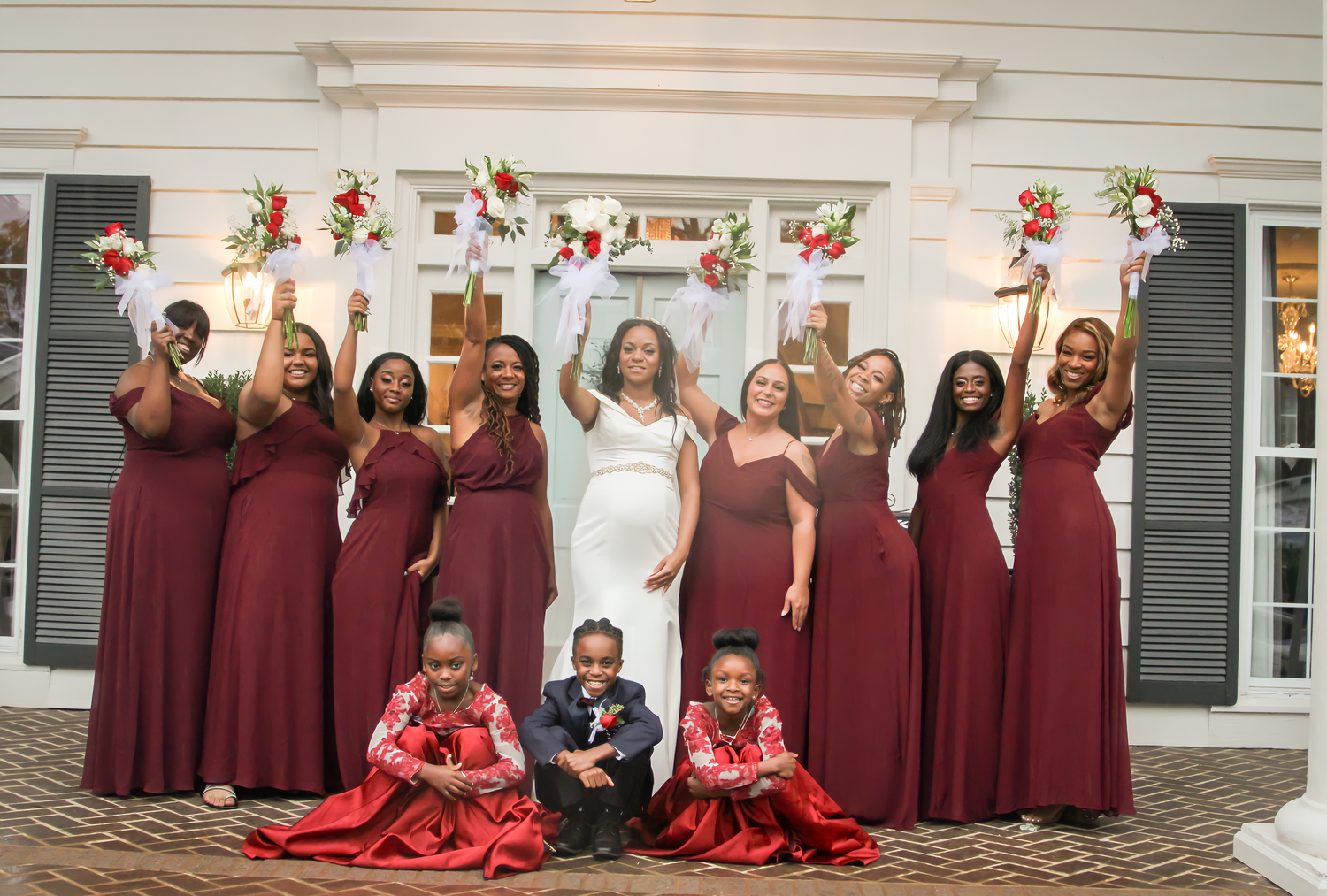 Wedding party in burgundy dresses celebrates together against white building exterior.
