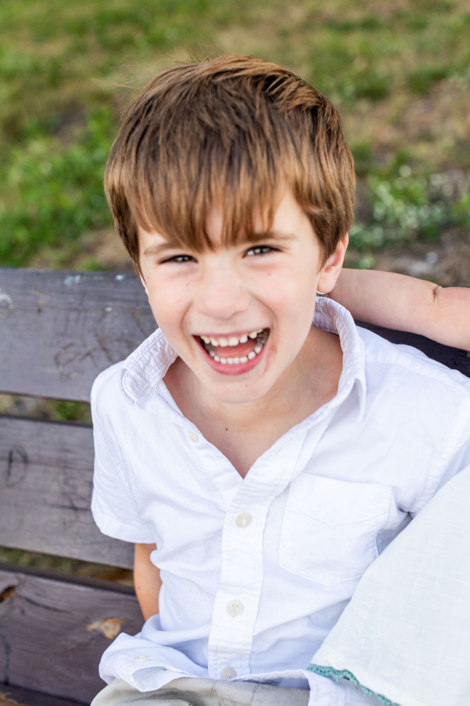 A cheerful young boy in a white shirt laughs joyfully while sitting on a wooden bench outdoors.