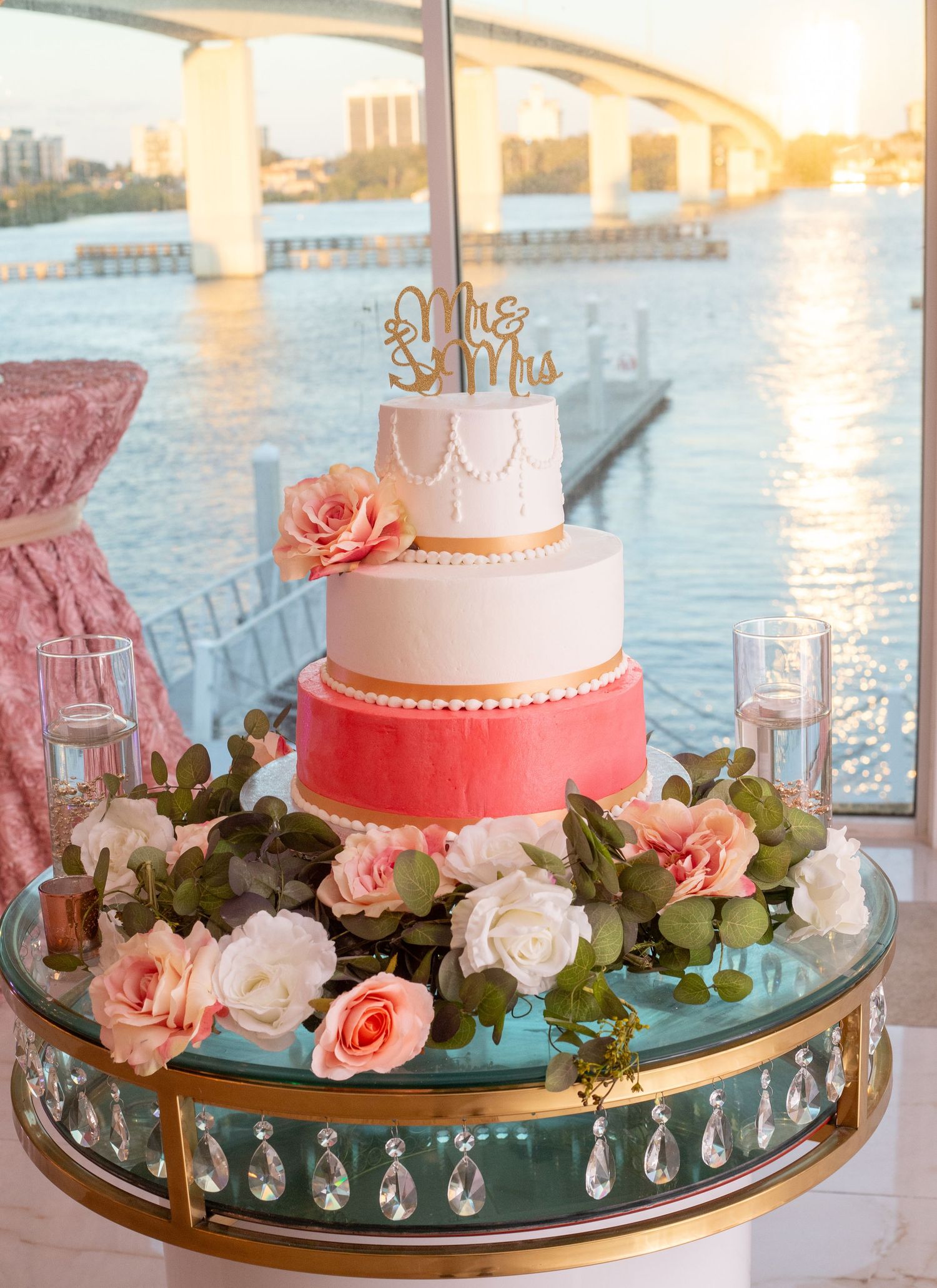 Two-tiered coral and white wedding cake adorned with roses on a crystal-trimmed stand by the water.