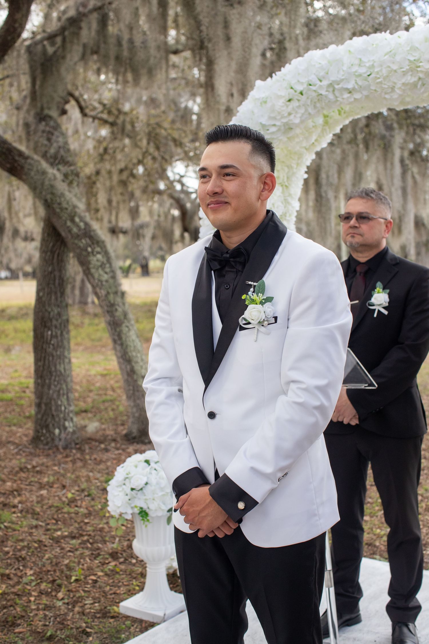 Groomsmen in white tuxedo jackets and black pants stand together during an outdoor wedding ceremony.