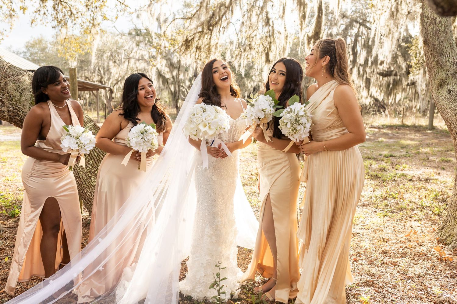 Bridesmaids in blush pink dresses pose together outdoors holding white bouquets.