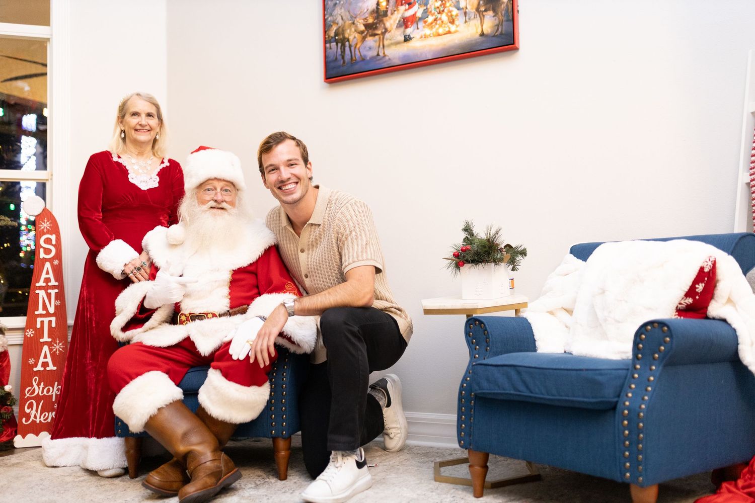 Holiday photo session with Santa Claus on blue chaise lounge in decorated room.