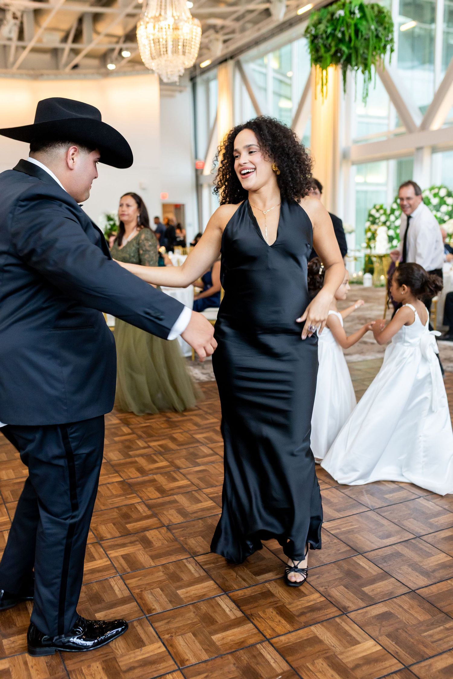 Couple dancing joyfully at a wedding reception, woman in black gown, man in black suit and cowboy hat.
