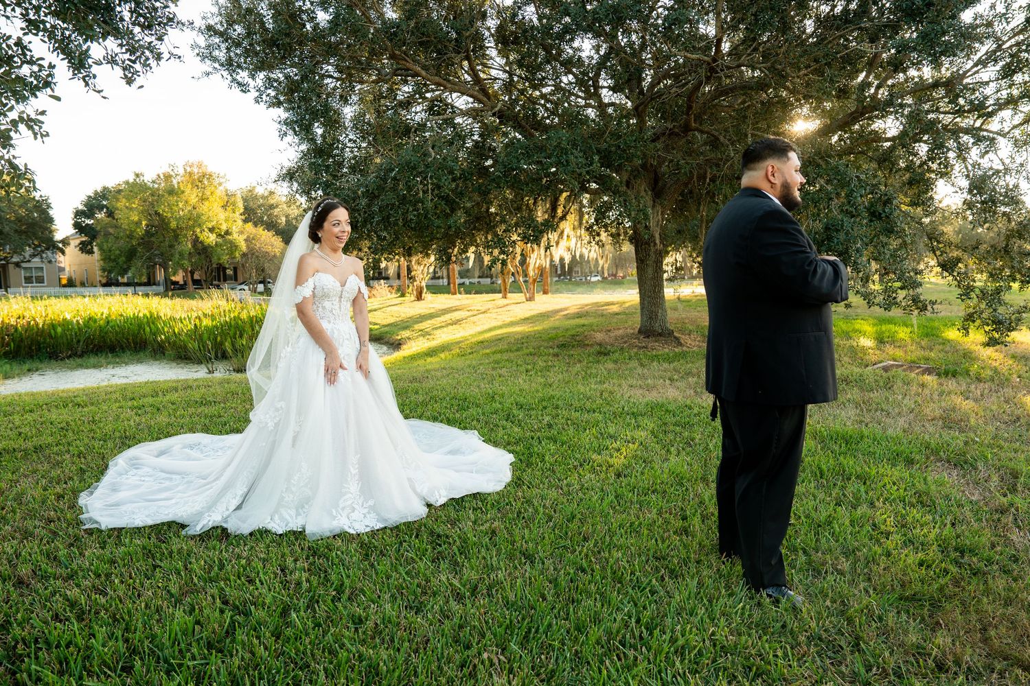 Bride in white ball gown with veil stands on green lawn as groom in black suit turns away for first look.