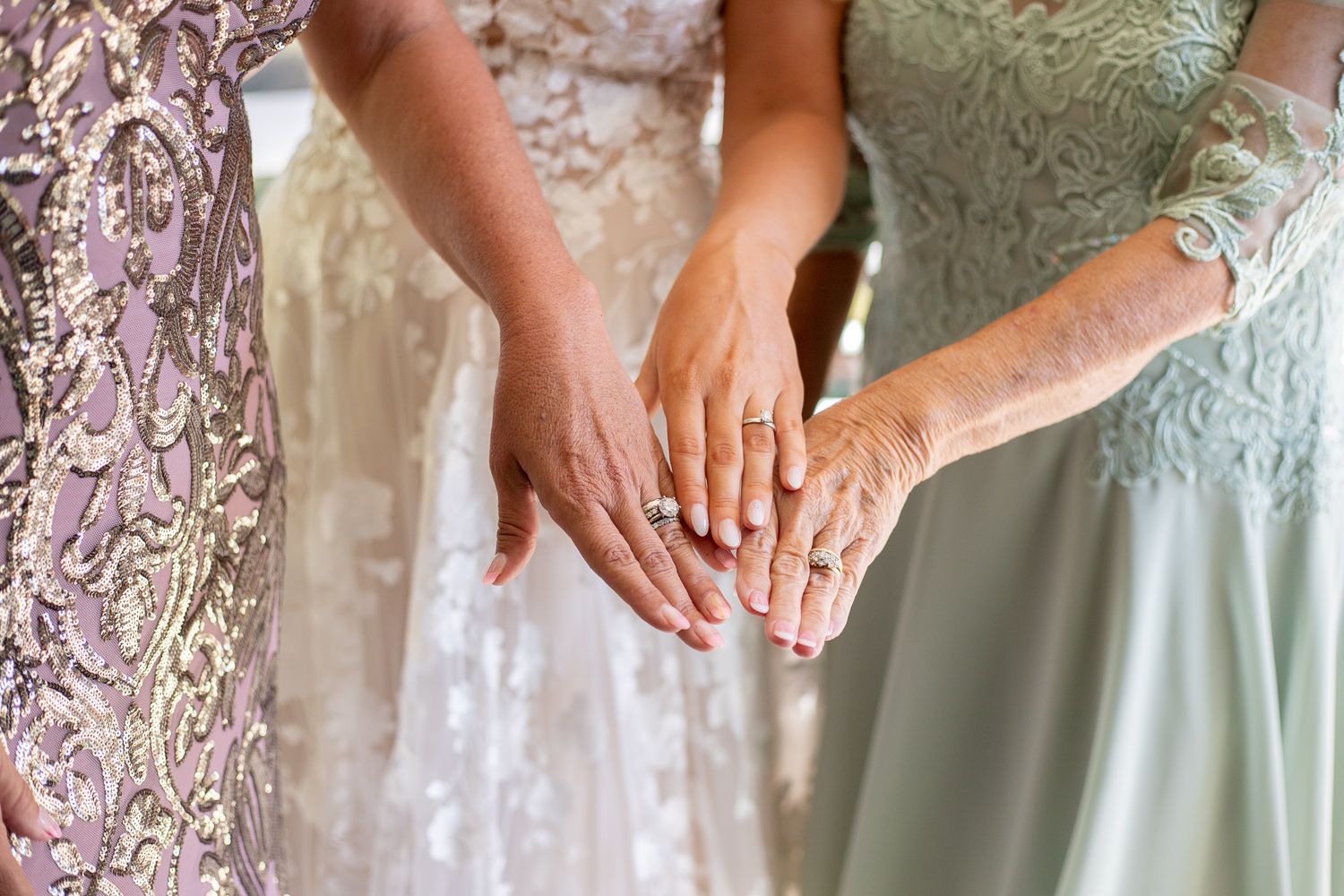 Three women in formal gowns display their hands together, showcasing a diamond engagement ring at a wedding celebration.