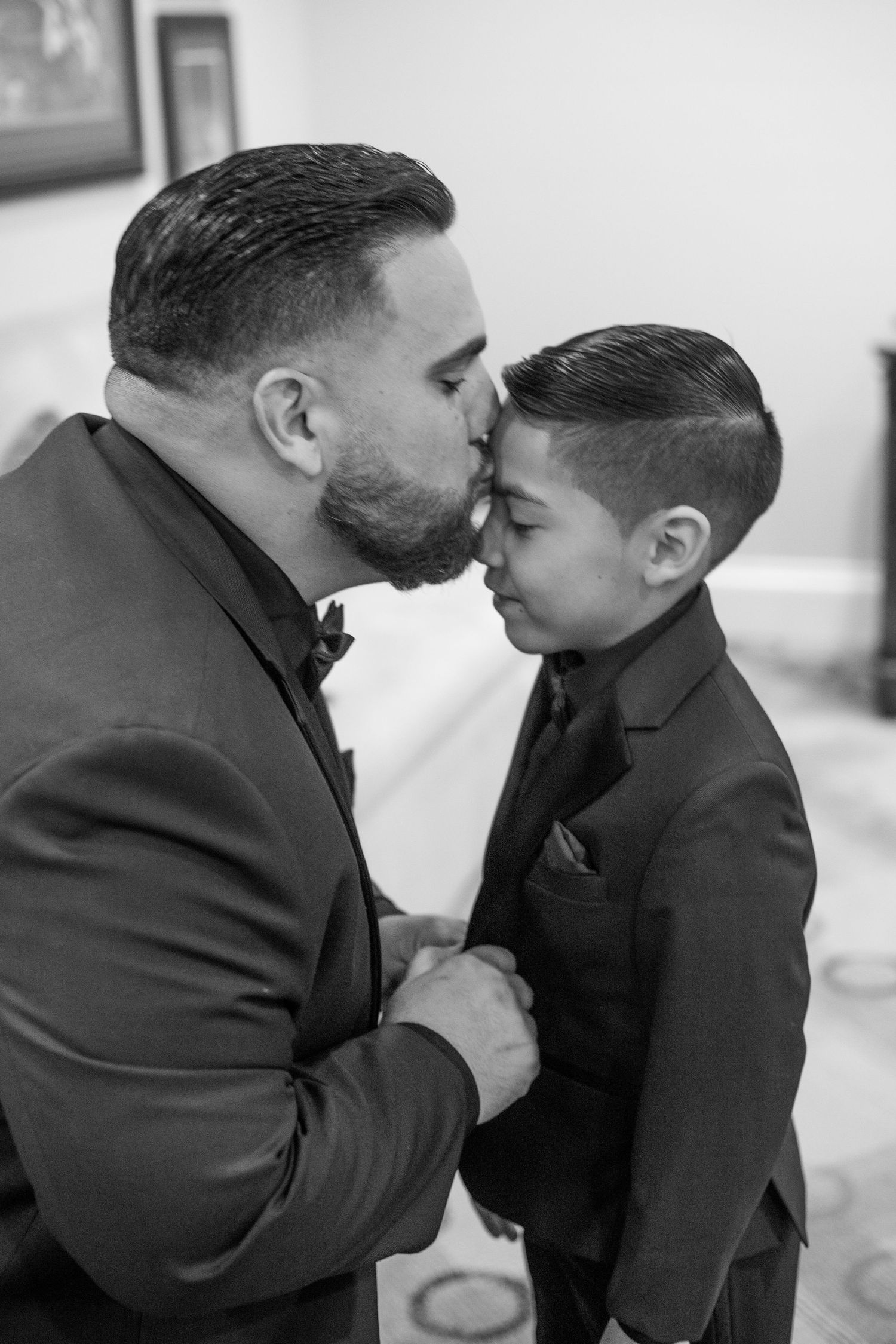 Black and white photo of a father in a suit kissing his young son on the forehead at a wedding.