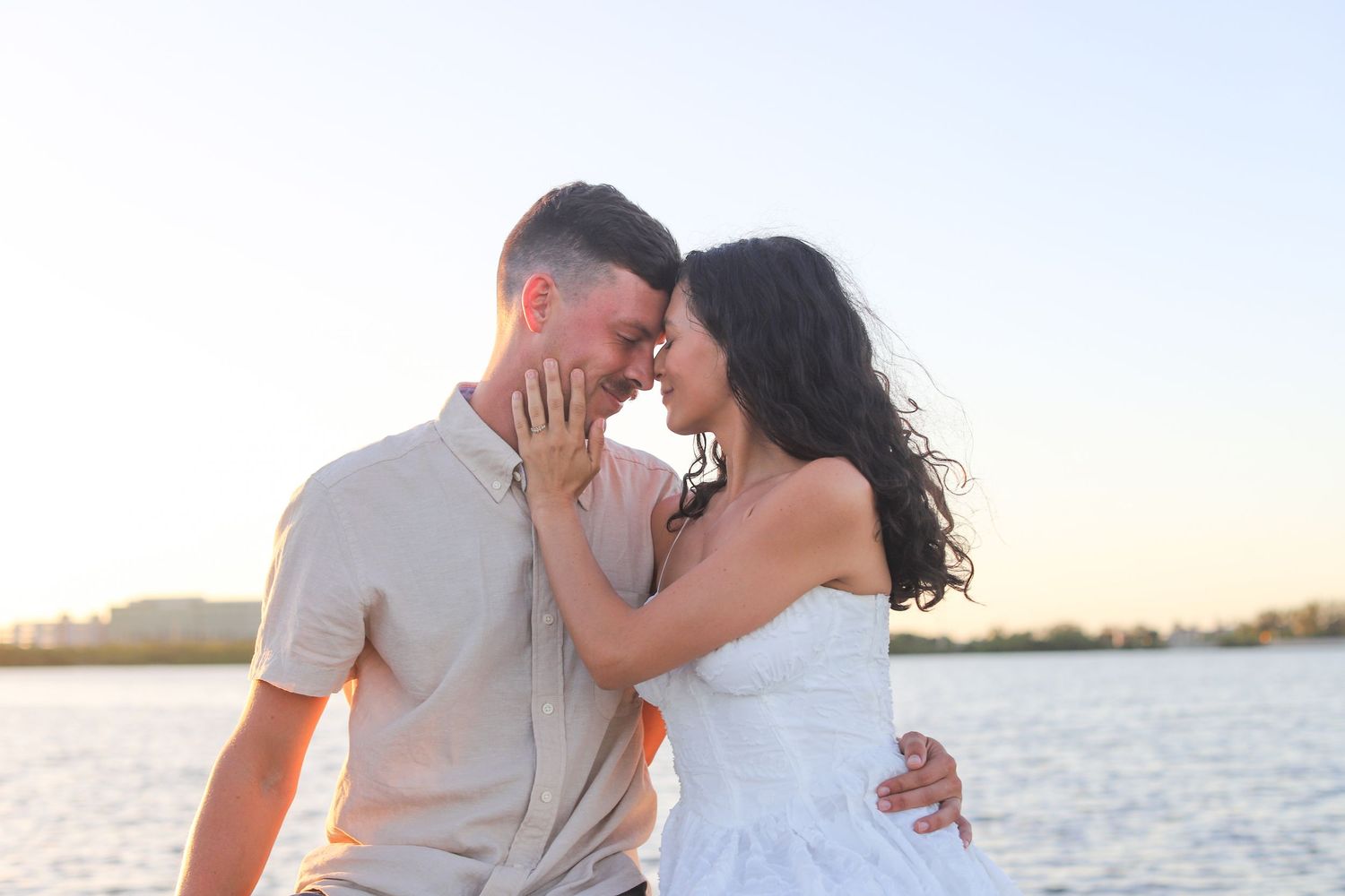Romantic couple sharing an intimate moment by the water at sunset, woman in white dress and man in beige shirt.