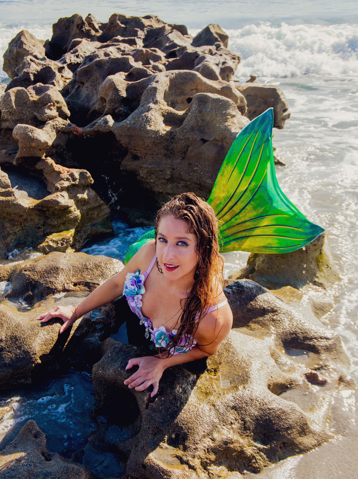 Costumed mermaid with green and yellow tail poses among ocean rocks.