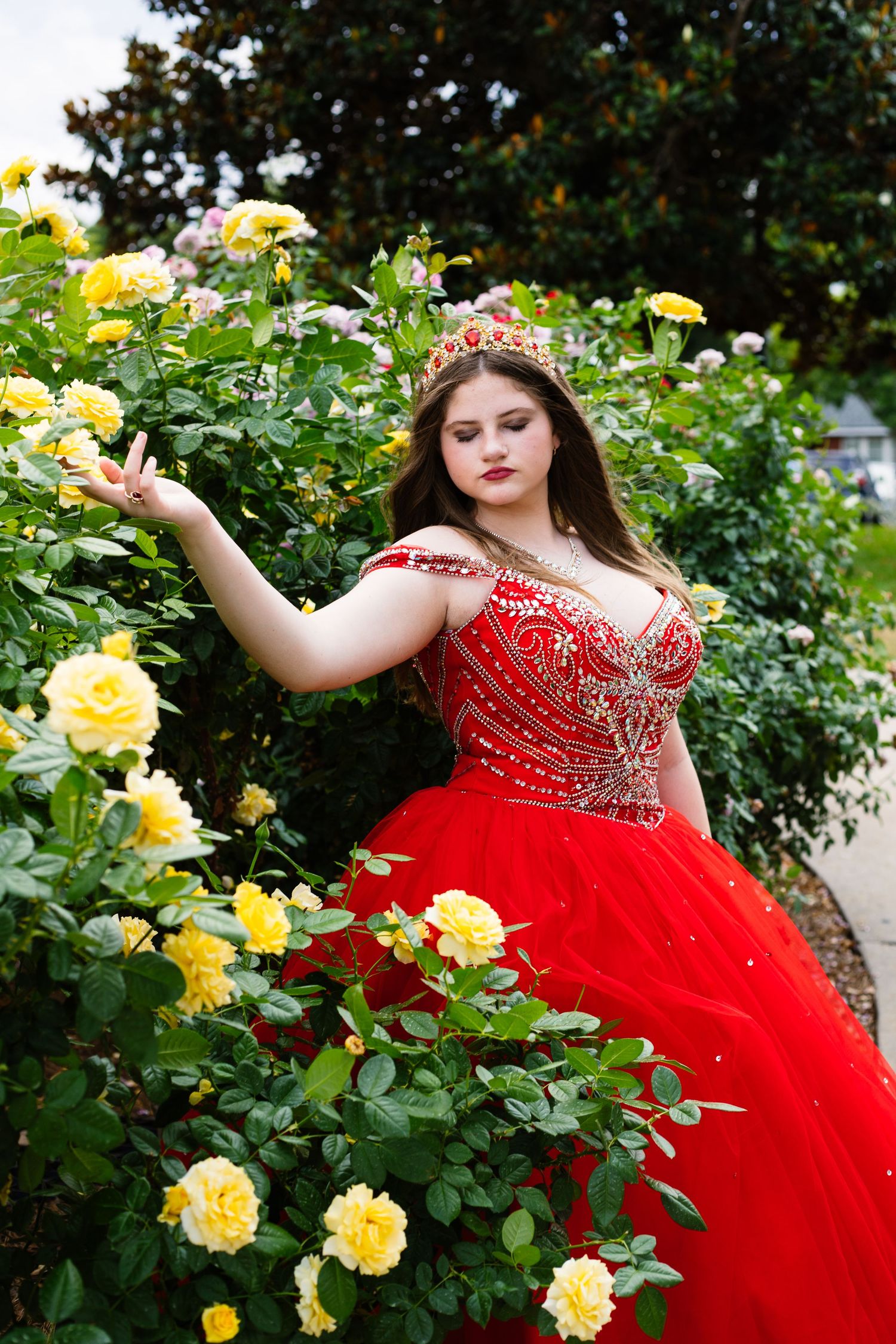 Elegant red ball gown photographed in a garden setting with blooming yellow roses.
