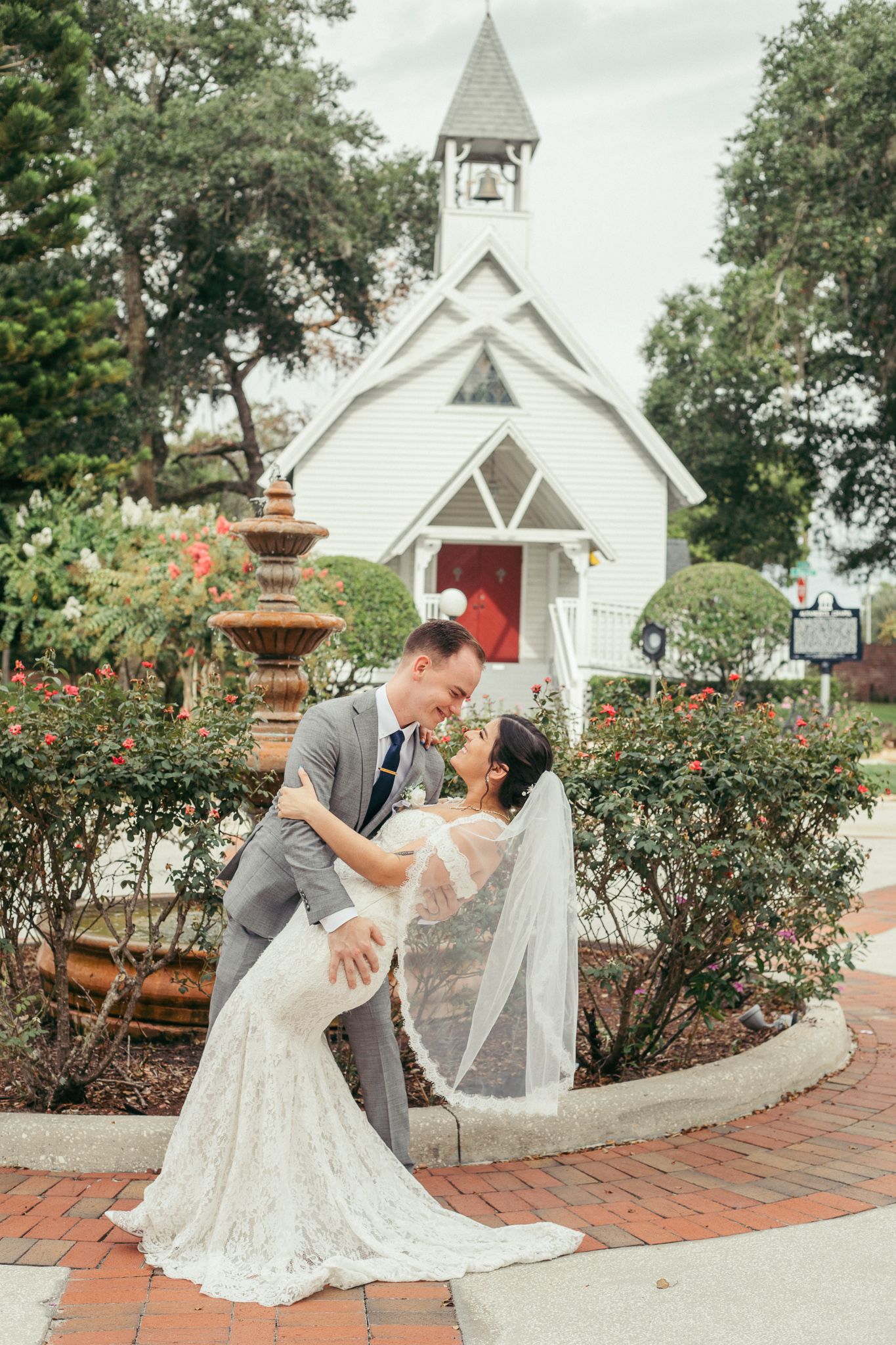 A romantic wedding scene in front of a white chapel with red roses and brick pathways.
