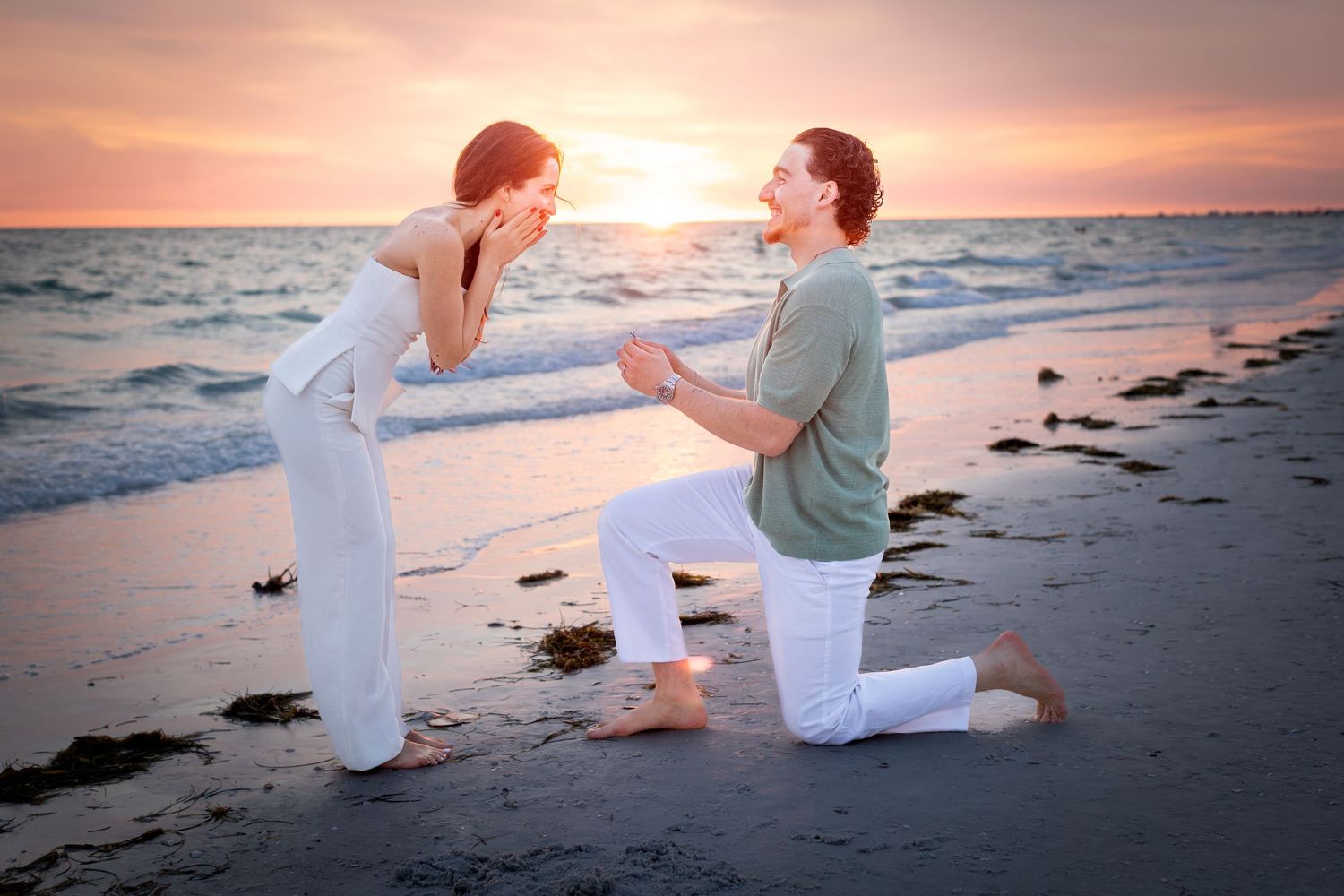 Man proposes to woman on a beach at sunset, kneeling in the sand as waves crash behind them.