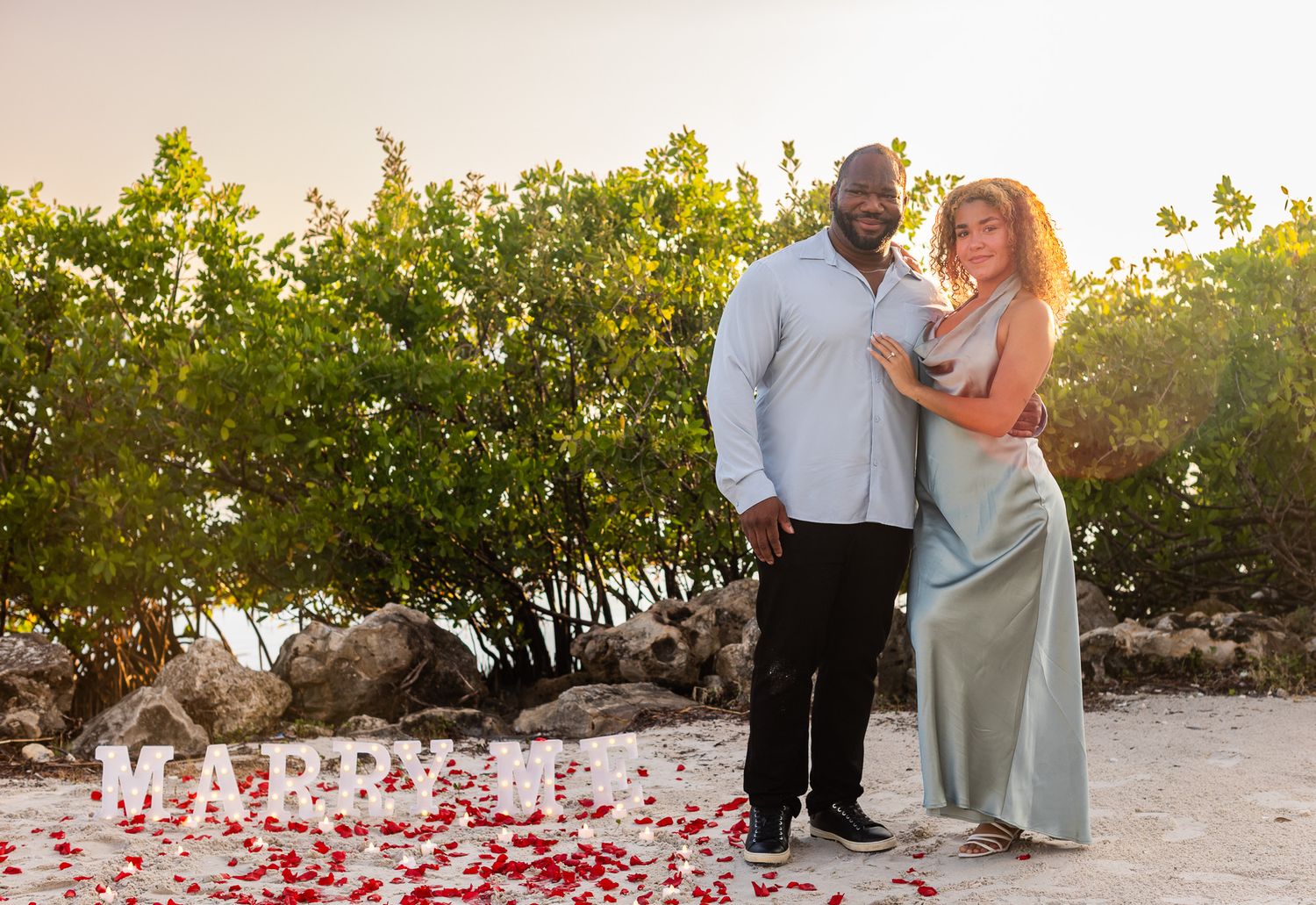 Couple embracing on tropical beach at sunset with rose petals and MARRY ME sign during romantic proposal.