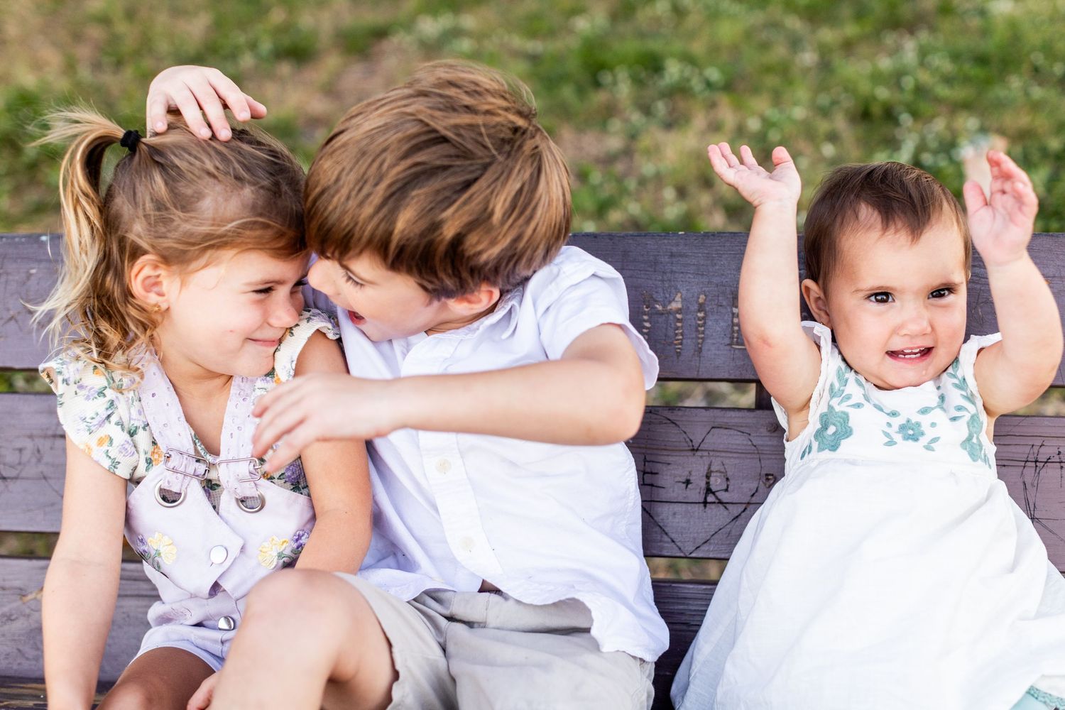 Three young children sitting on a wooden bench outdoors, two older kids hugging while a baby raises arms happily.