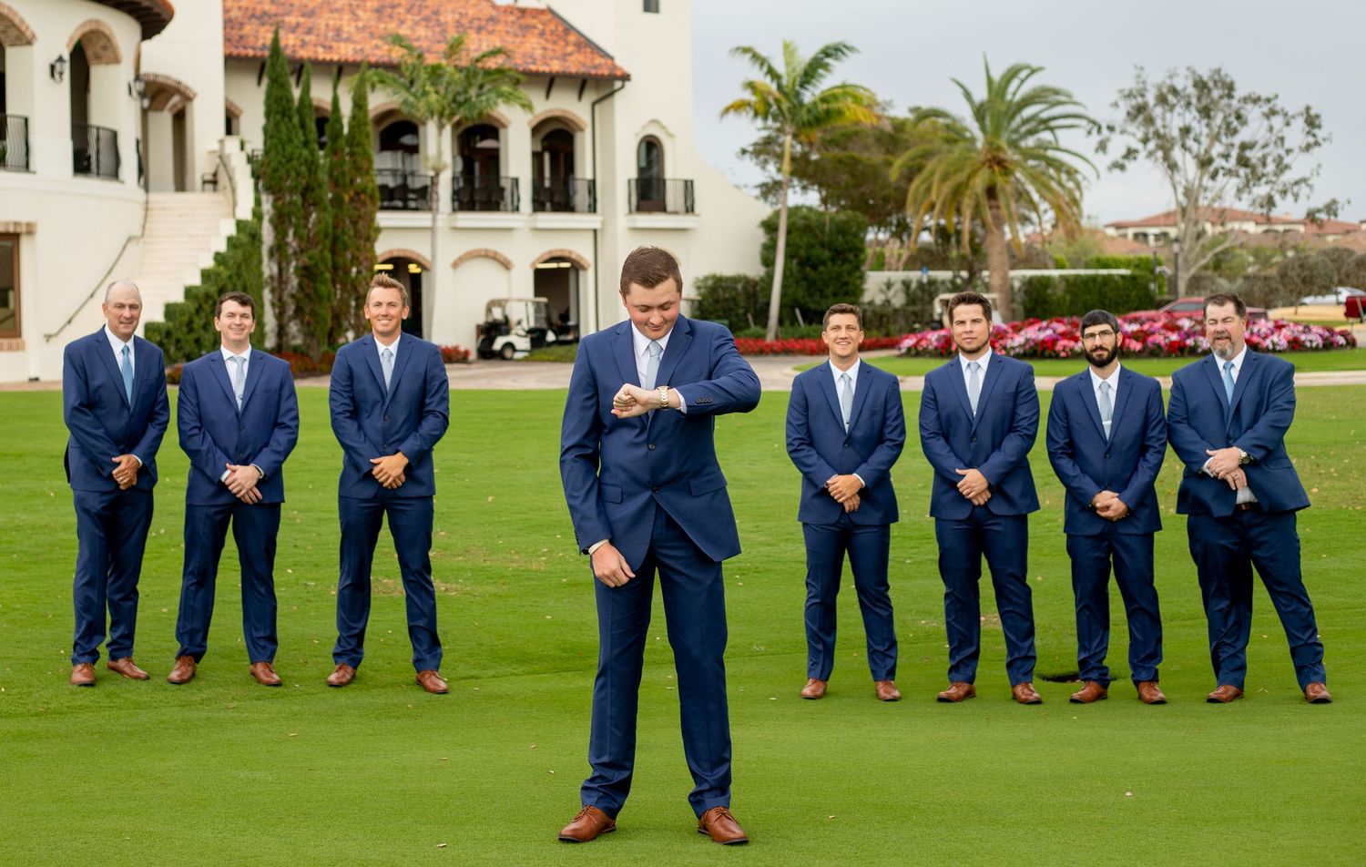 Groom checks watch in navy suit on golf course, with groomsmen lined up behind at elegant wedding venue.