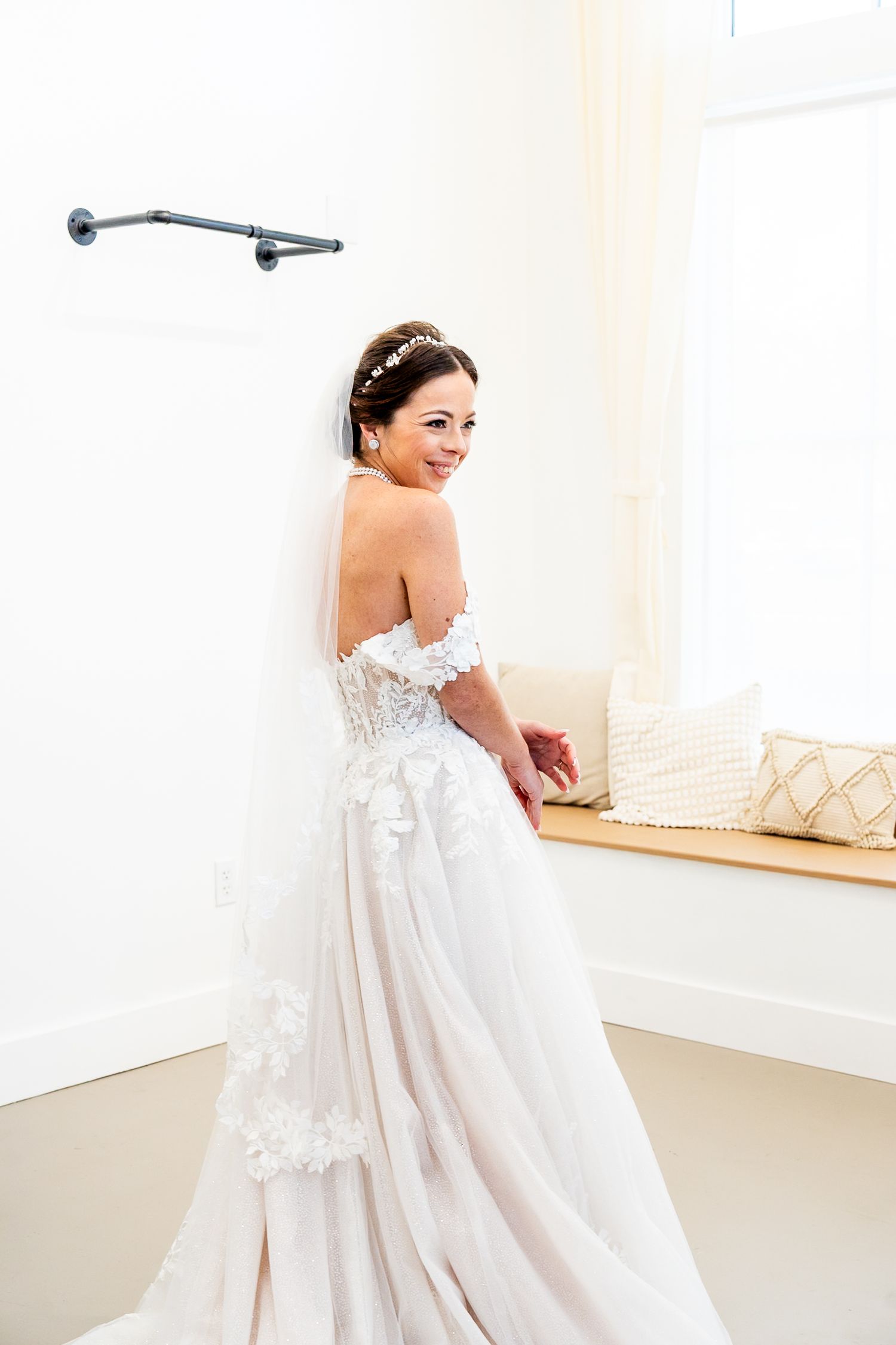 Bride in strapless floral lace ballgown with veil smiles over shoulder in bright white room.