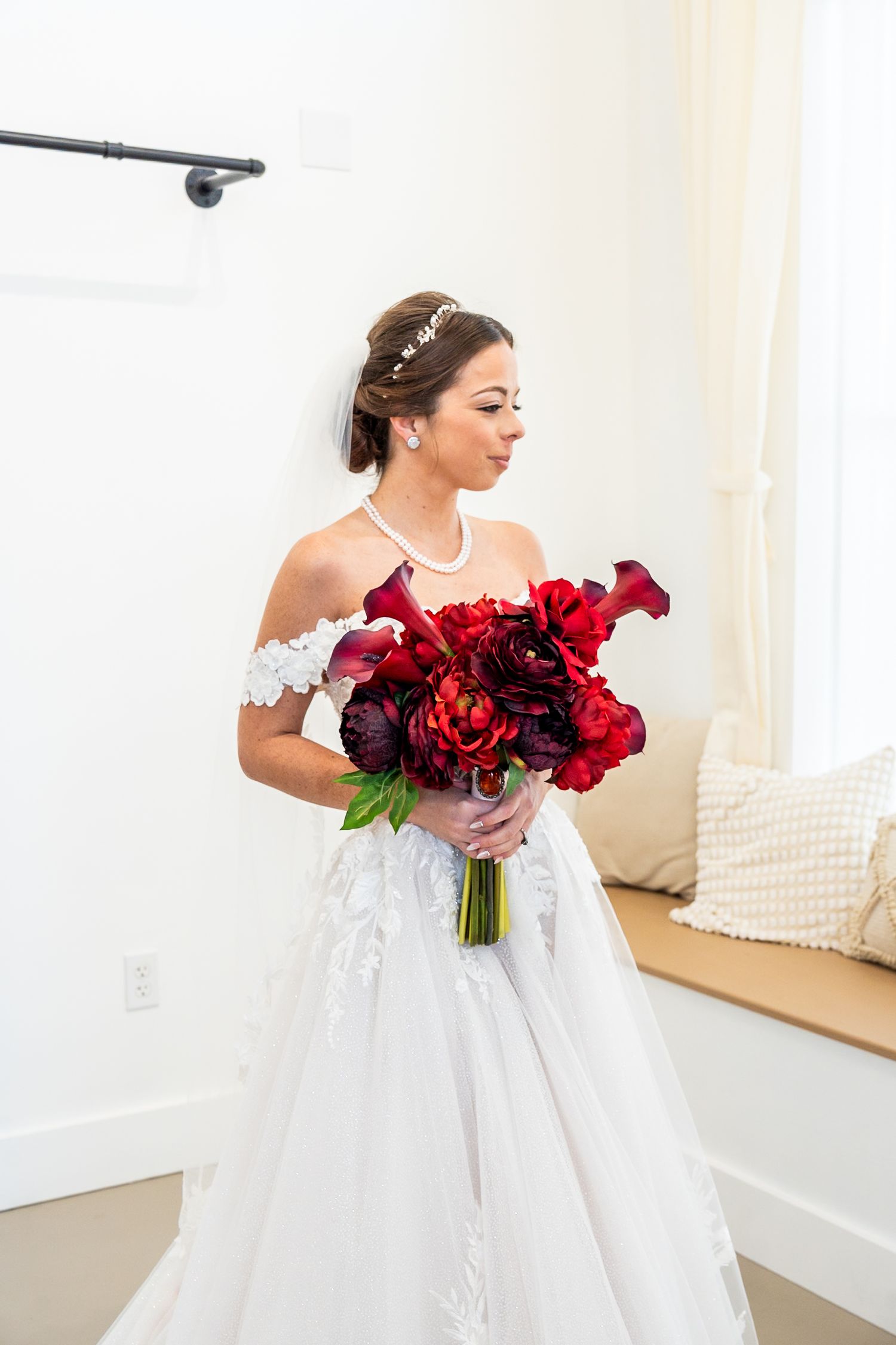 Bride in white off-shoulder gown holding red floral bouquet with calla lilies and peonies, wearing pearl necklace and hair piece.