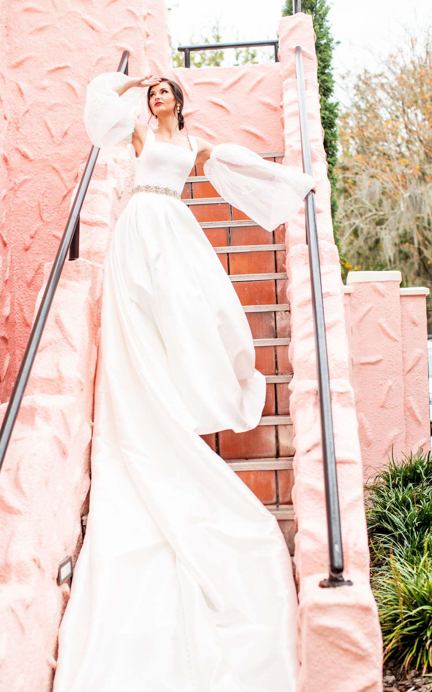 Bride in flowing white gown with bell sleeves poses on pink staircase outdoors.