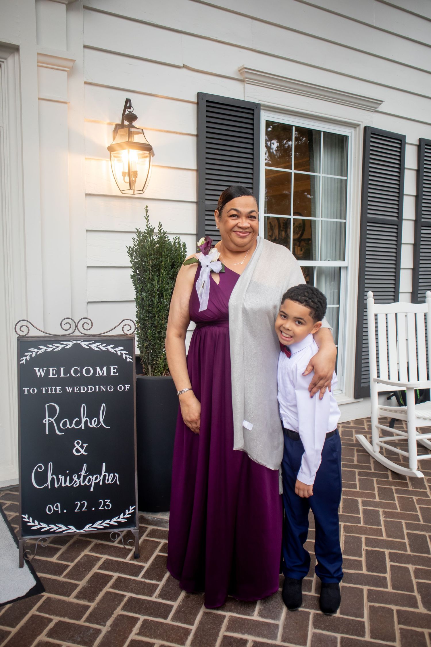 Woman in purple gown and boy in suit pose by Rachel and Christopher wedding welcome sign at elegant venue.