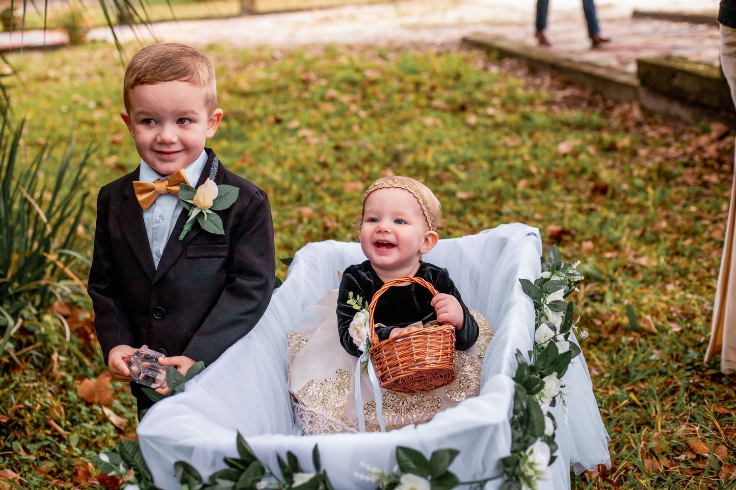 Two young boys in formal wedding attire, one as ring bearer and one as flower girl, smiling outdoors at a wedding ceremony.