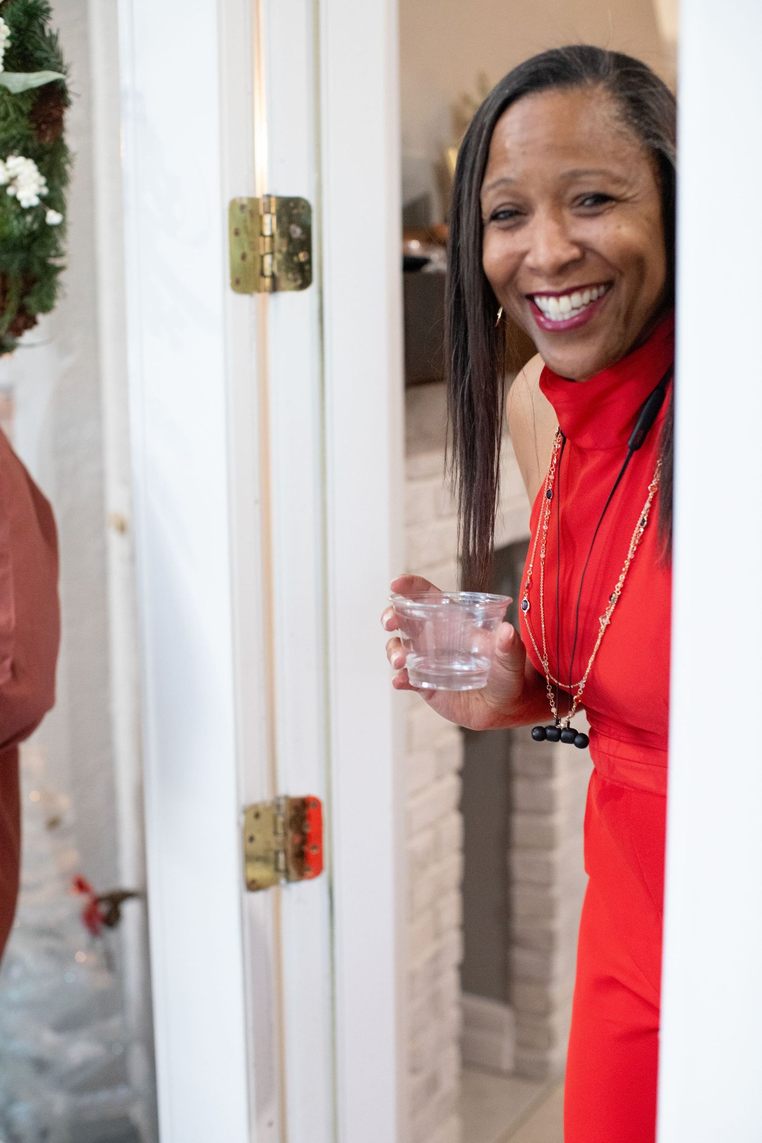 Person in bright red dress peeking through white doorway with a glass.