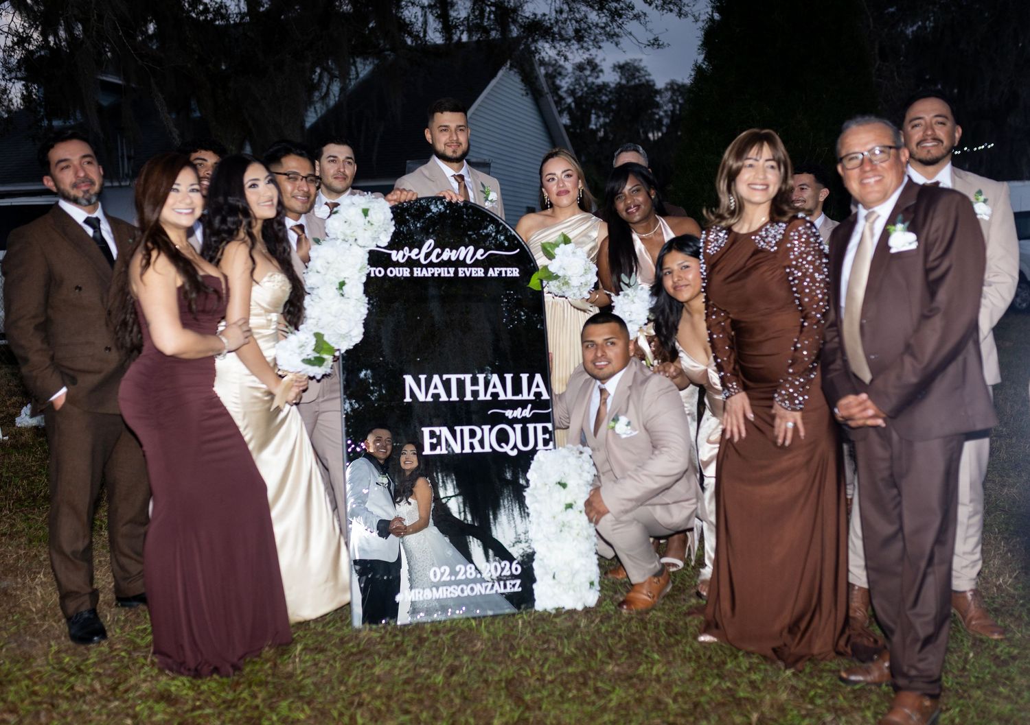 Wedding party members in burgundy and brown attire pose together by a welcome sign.