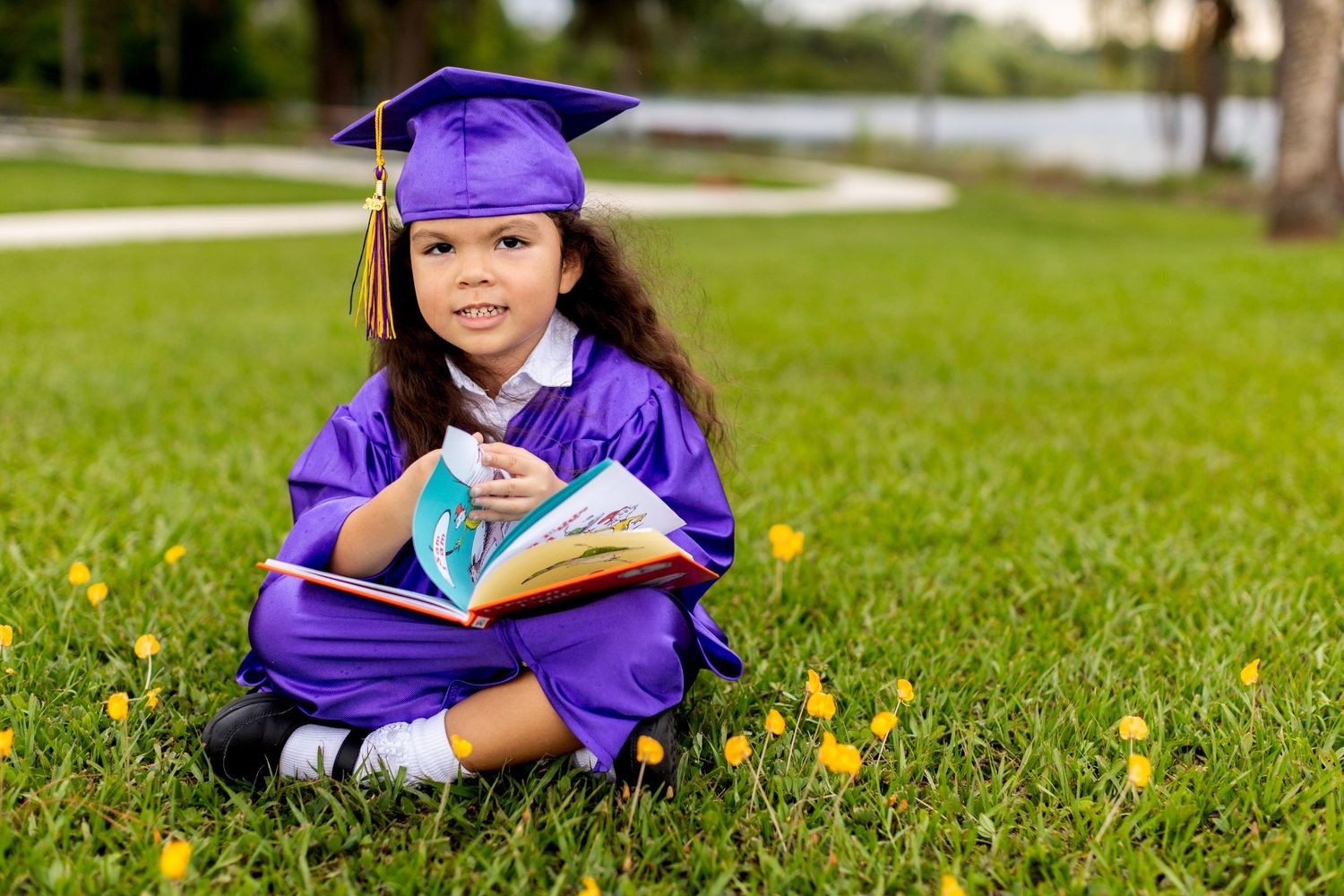 Graduate in purple cap and gown sits cross-legged on grass with yellow flowers holding a book.