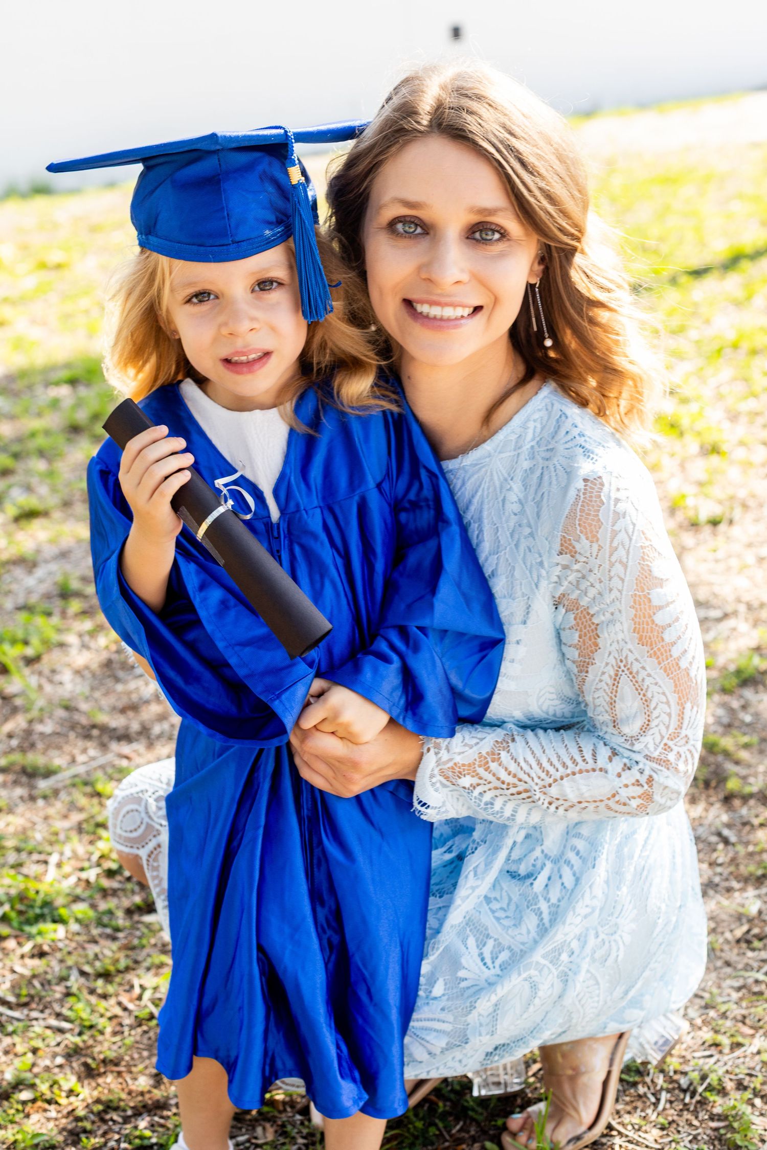 A graduation celebration photo featuring a small graduate in blue cap and gown with a loved one in a lace dress outdoors.