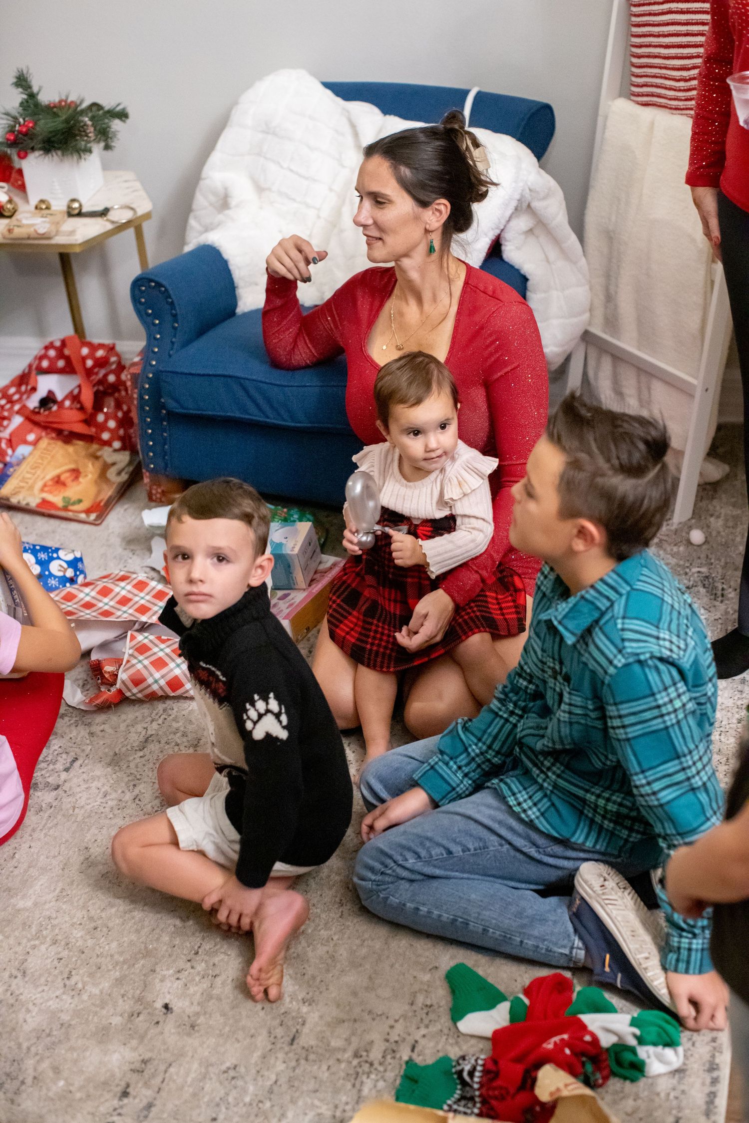 A holiday gathering shows people sitting on the floor opening presents near a blue armchair.