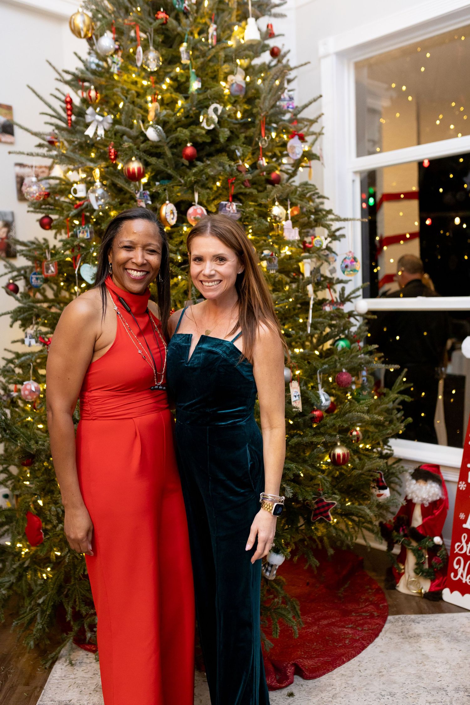 Two friends in formal dresses smile together in front of a decorated Christmas tree.